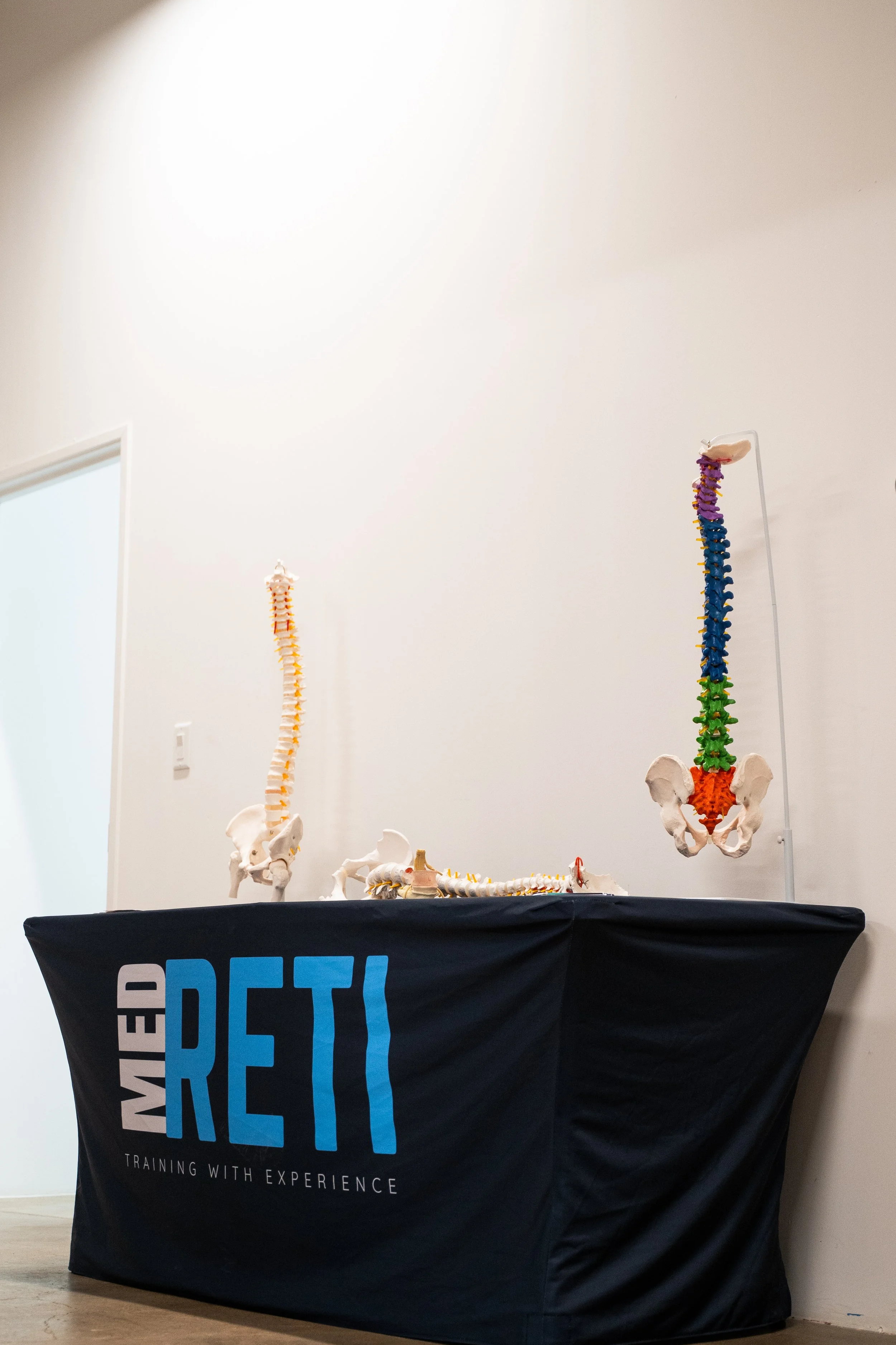 Educational display of human spine and pelvis models on a table with a black cloth that reads 'MED RETI TRAINING WITH EXPERIENCE' in a room with white walls.