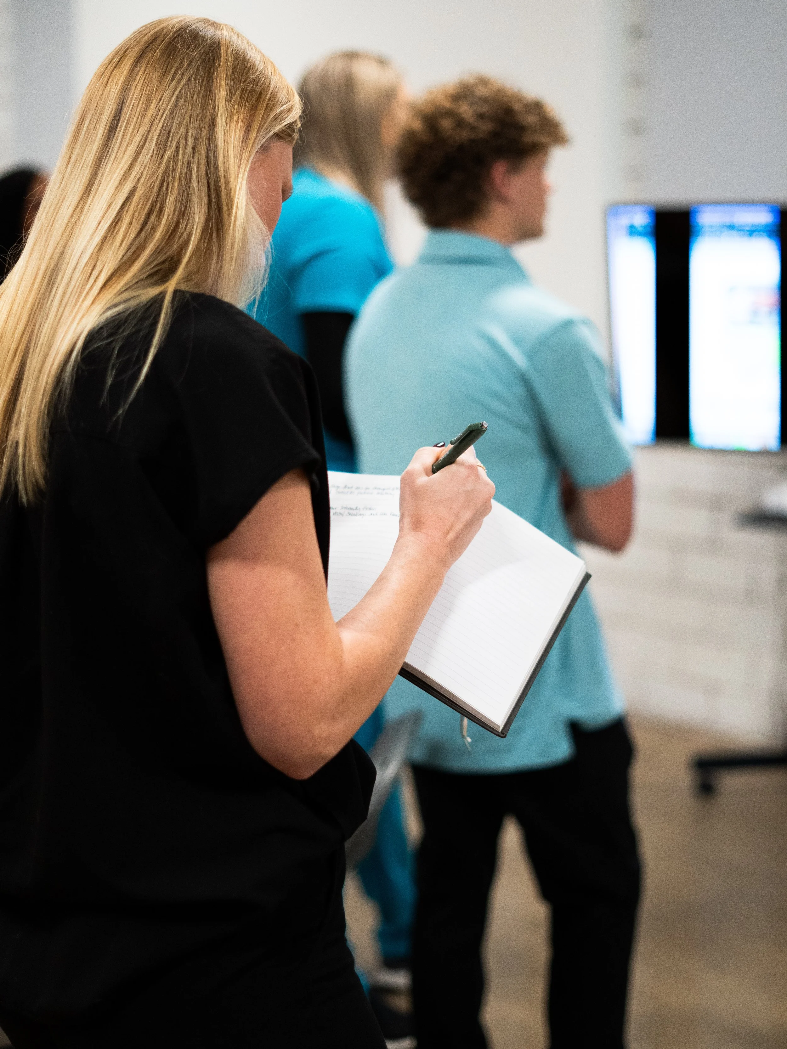 A woman taking notes while standing in line at a tech event, with two people in front of her looking at screens.
