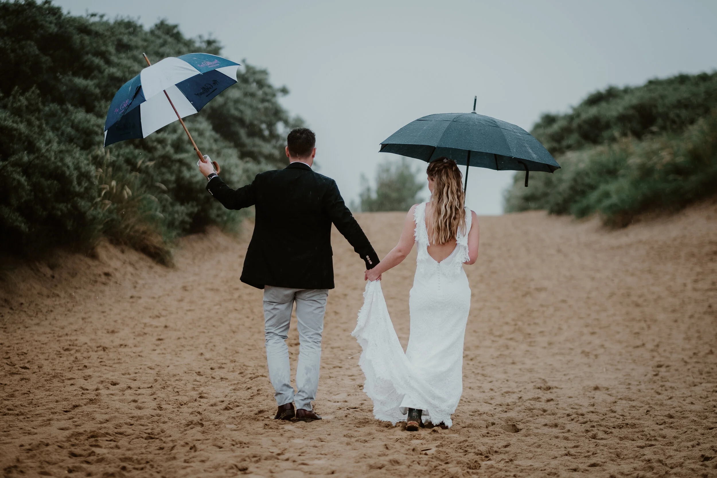 A couple dressed in wedding attire walk hand in hand on Camber sands beach, holding umbrellas on a cloudy day.