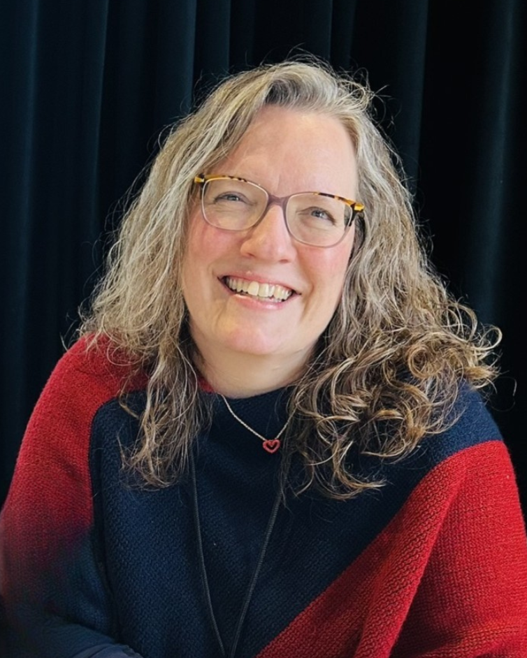 A woman with glasses and curly gray hair smiling and holding a blue puzzle piece in front of a gray display board with colorful papers. She is wearing a red cardigan and a black top, with signs that read "RADICAL WELCOME" and others behind her.