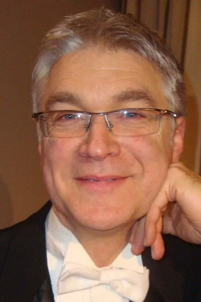 A middle-aged man with gray hair and glasses, dressed in a tuxedo with a white bow tie, smiling at the camera, indoors.