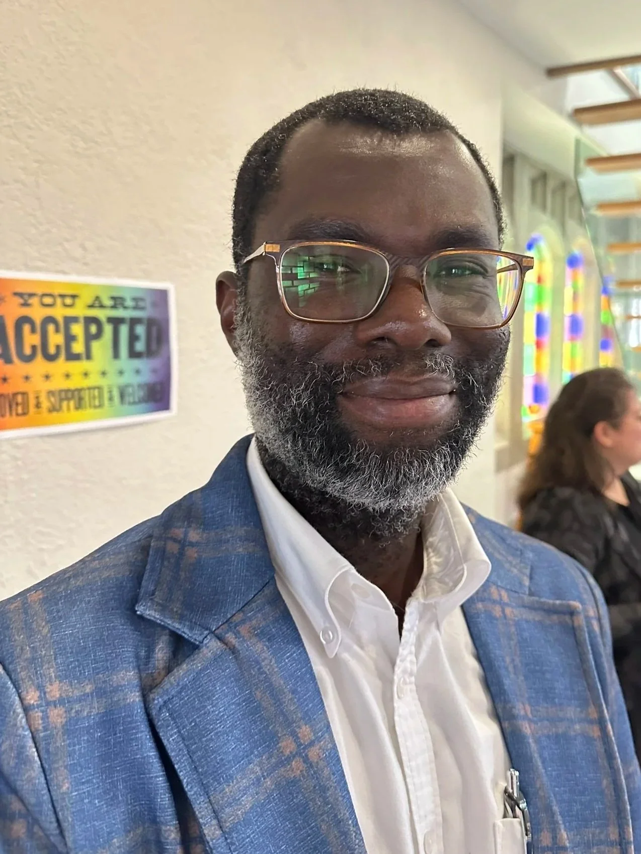 A man with glasses and a beard wearing a colorful, patterned shirt in front of a brick wall.