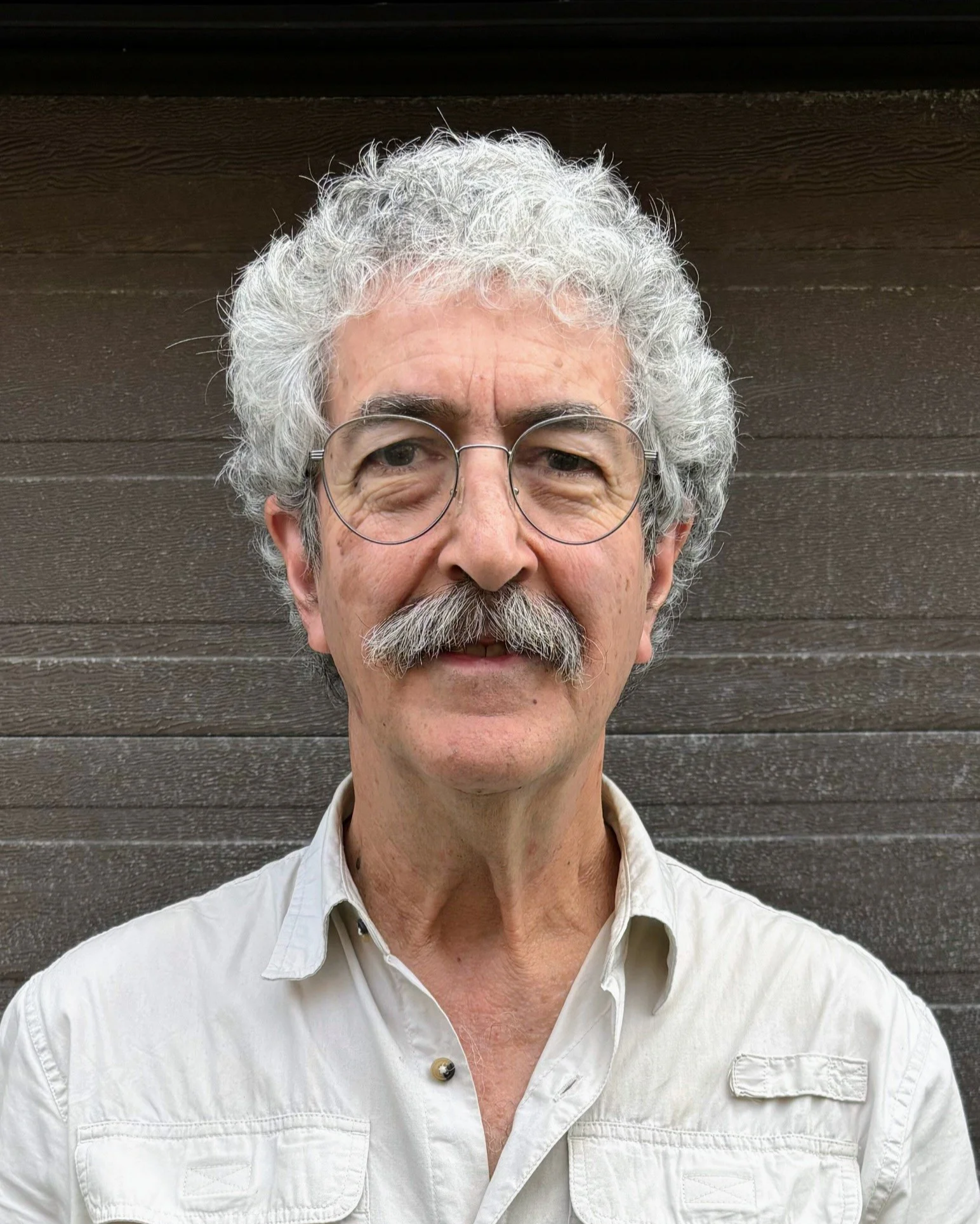 A man with curly gray hair, glasses, and a mustache, wearing a light-colored button-down shirt, standing in front of a wooden background.