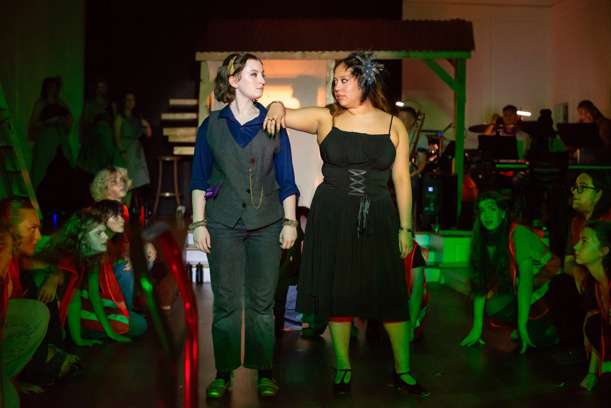 Two actors are looking at each other intensely as the ensemble looks on behind then. The actor on the left is wearing a grey vest and pant suit with a purple button down. The actor on the right is wearing a strappy black dress with a corset and glitt