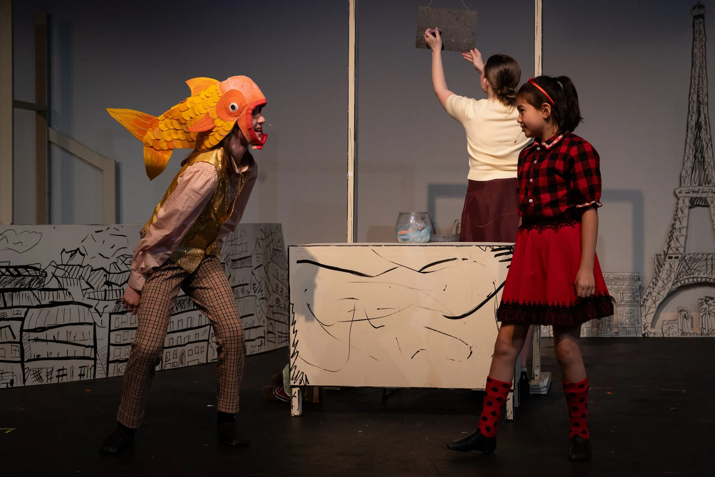A child wearing an orange fish hat is leaning in towards another child wearing a red plaid top and red skirt. 