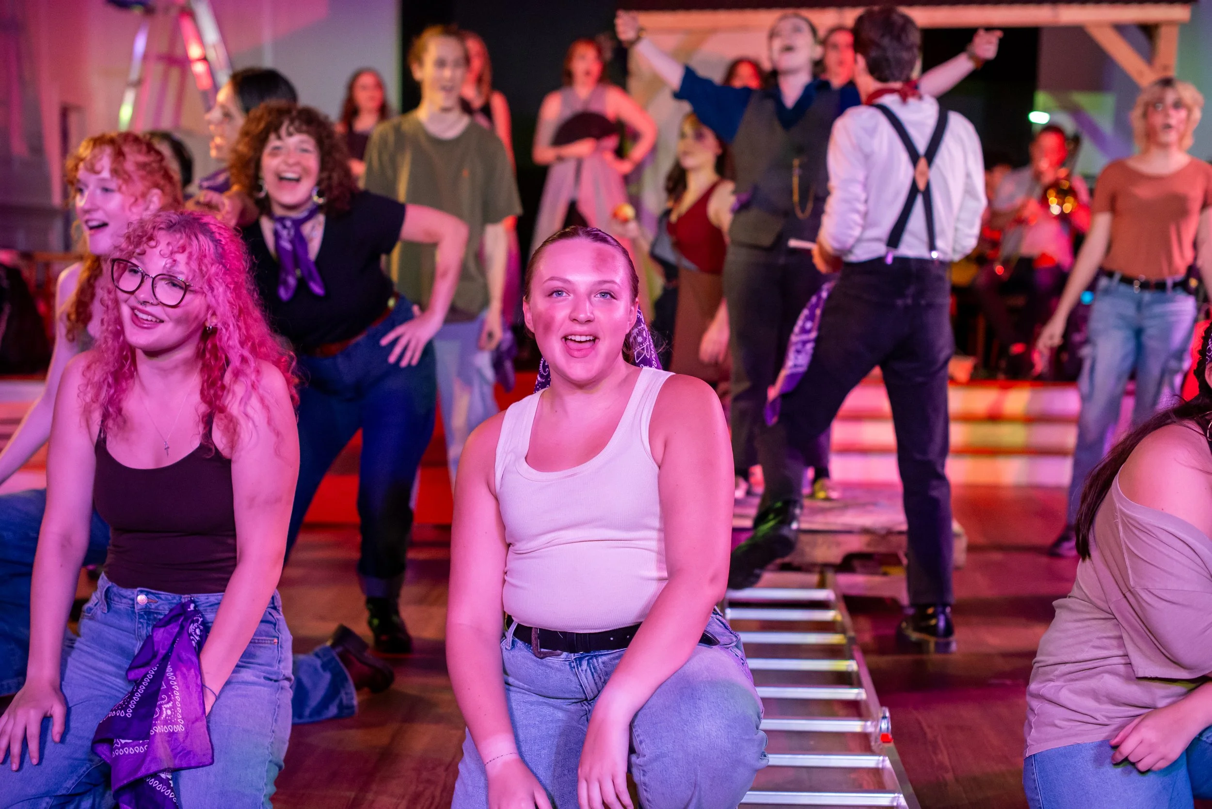 The ensemble of Hadestown are positioned in a stage picture. Two young actresses are kneeled facing the audience and singing. They are wearing tank tops with jeans, and are covered in dirt and grime. 