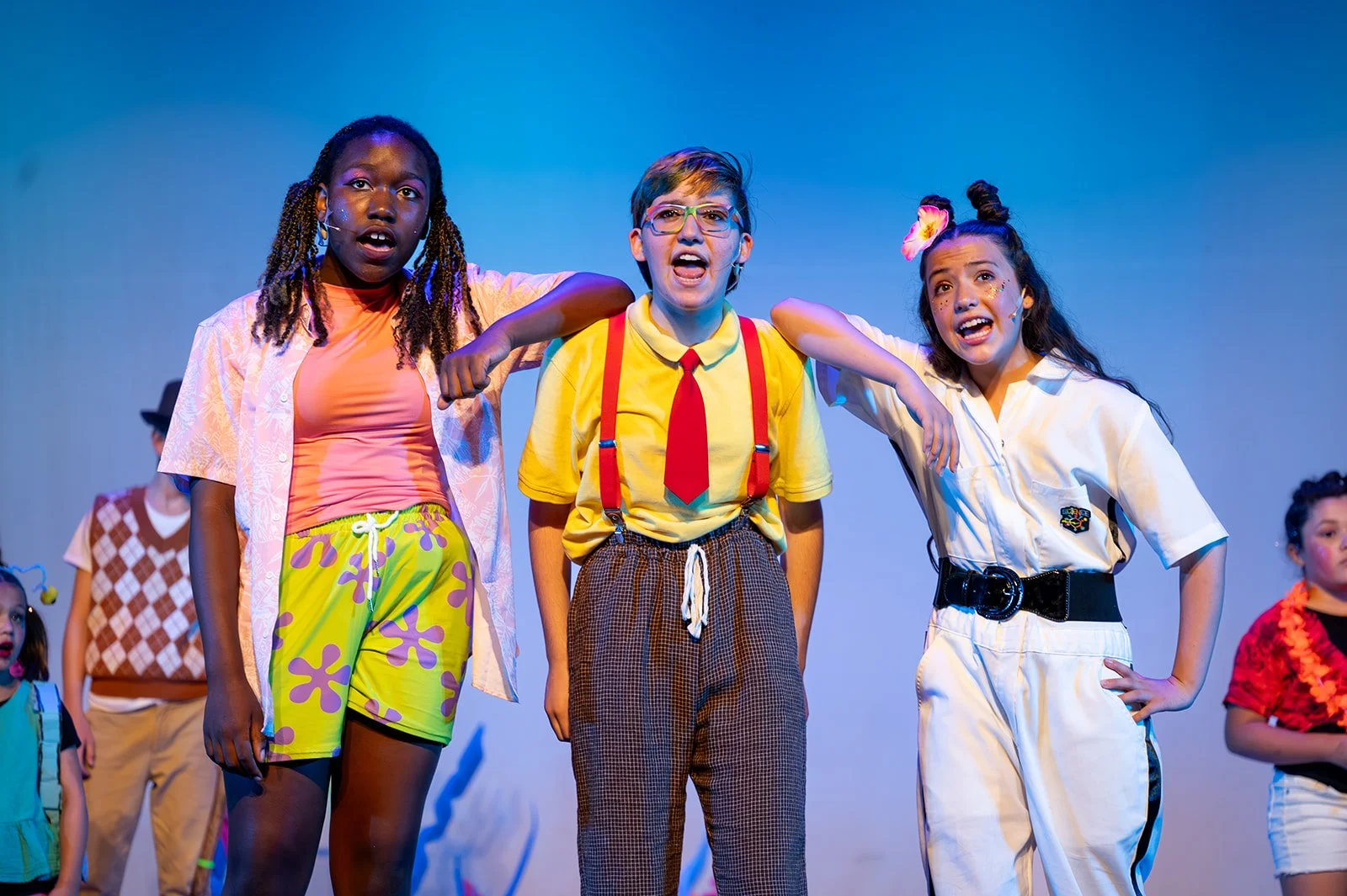 Three actors playing Patrick, Spongebob, and Sandy Cheeks are pictured. Patrick is wearing green and purple board shorts with a pink top and pigtails. Spongebob is wearing a yellow polo with a red tie and brown pants. Sandy Cheeks is wearing a white 