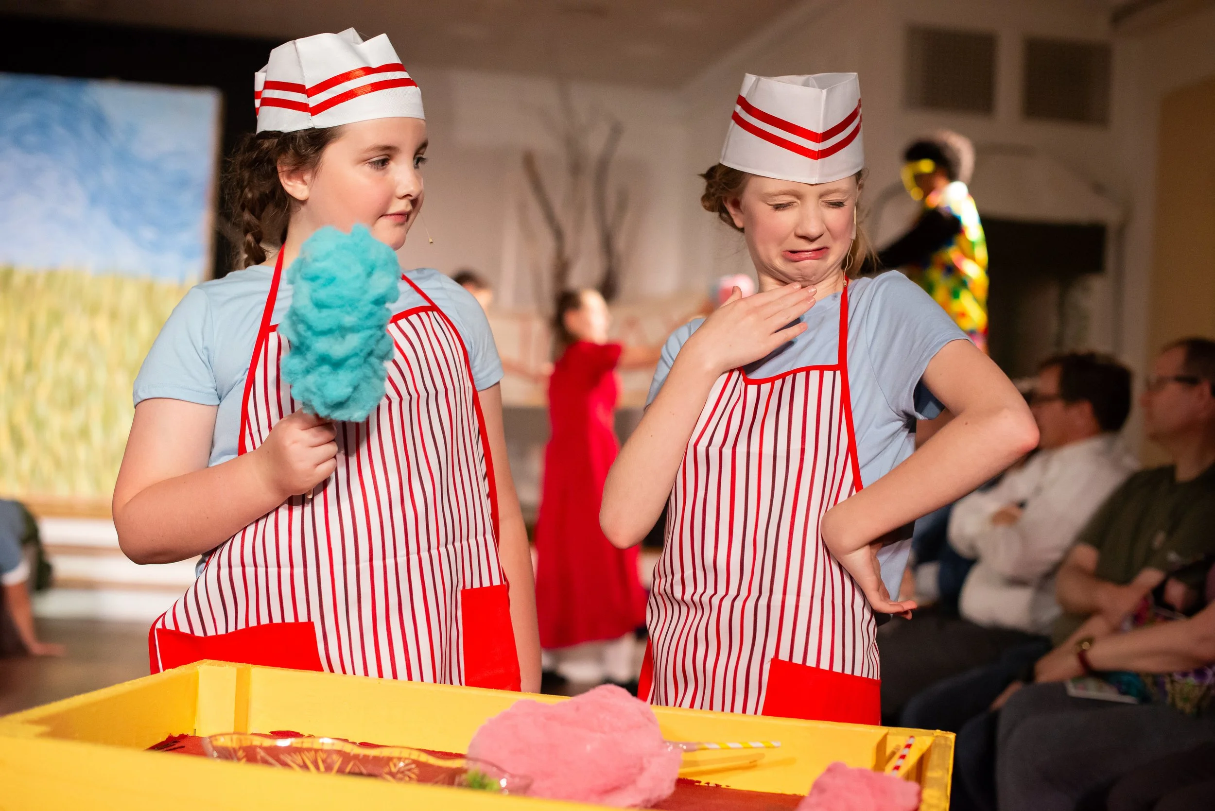 Two young ladies dressed as concessions workers are having a negative reaction to their cotton candy. They are wearing striped aprons with blue tshirts underneath. 