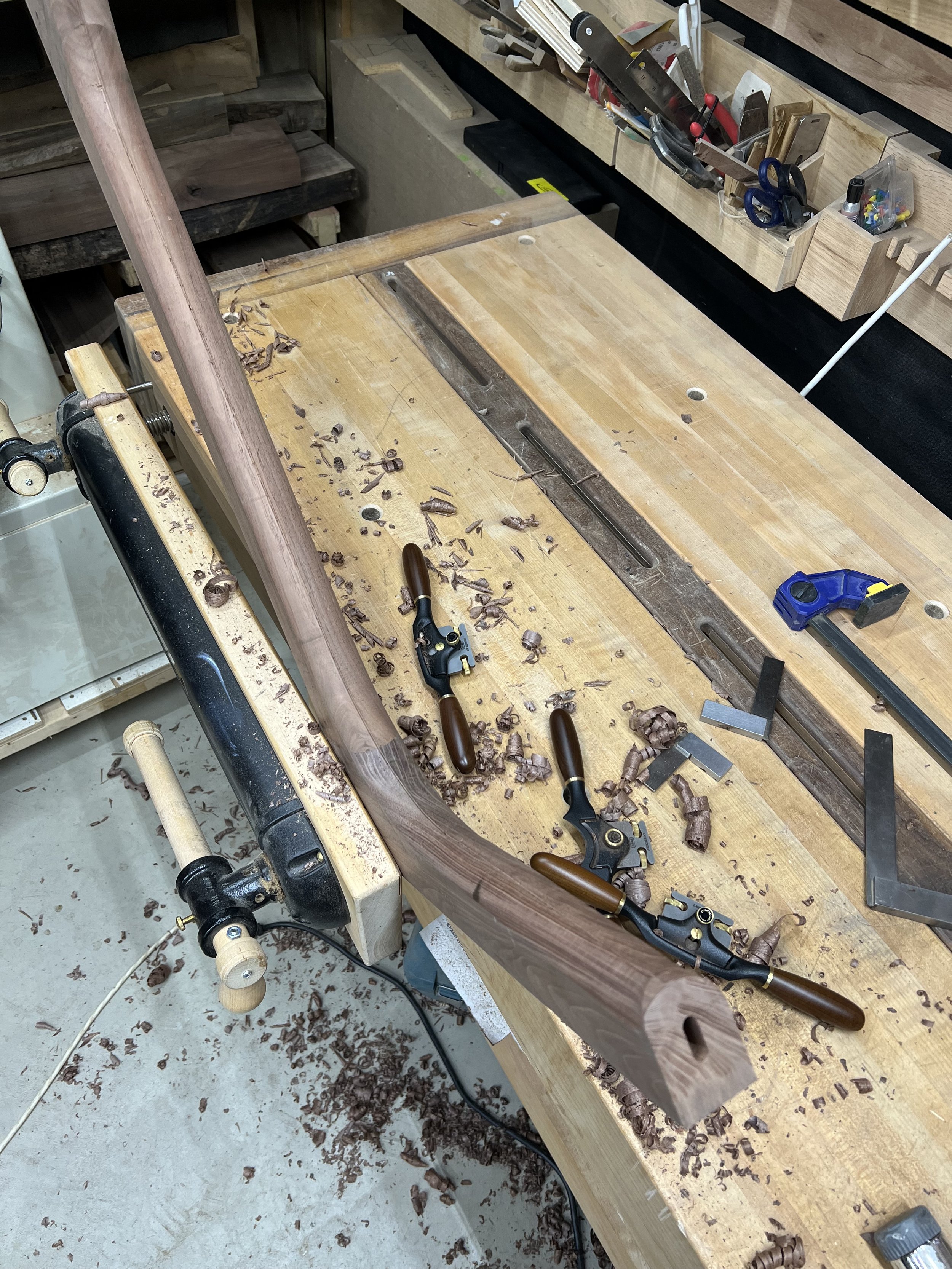 A woodworking workshop workbench with wood shavings, carving tools, and clamps. There are various woodworking tools and supplies organized on a wall-mounted shelf.