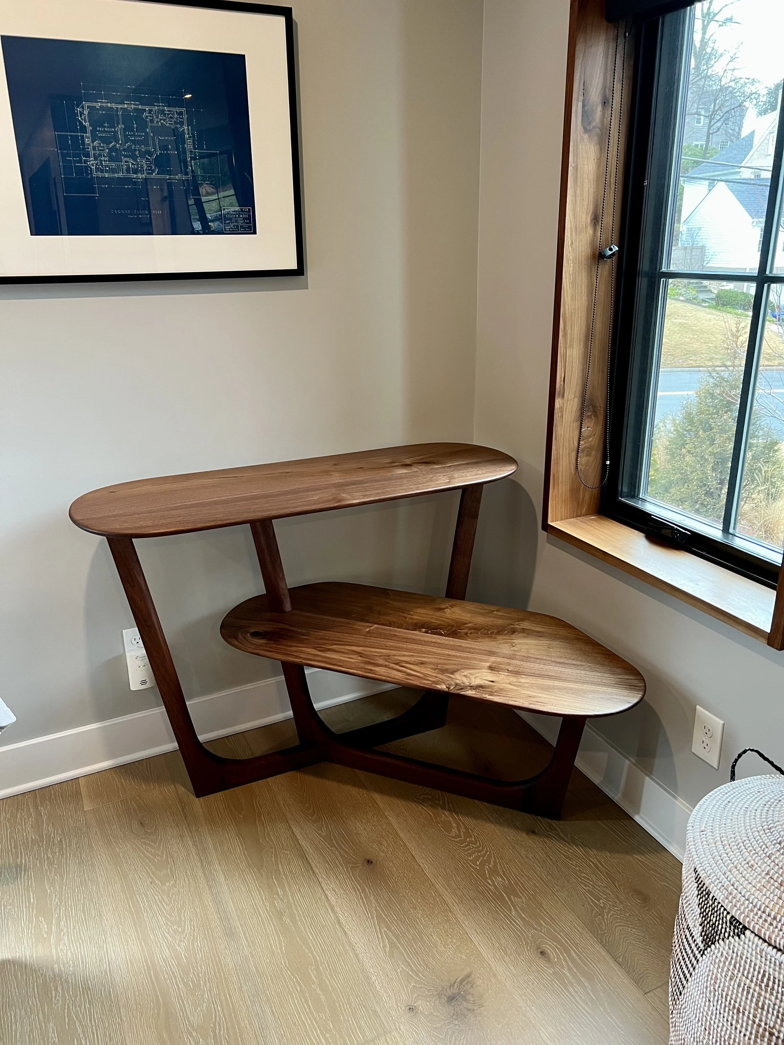 A wooden corner shelf with two levels, placed near a window with a wooden frame, in a room with light-colored walls and wood flooring. A framed blue print hangs on the wall.