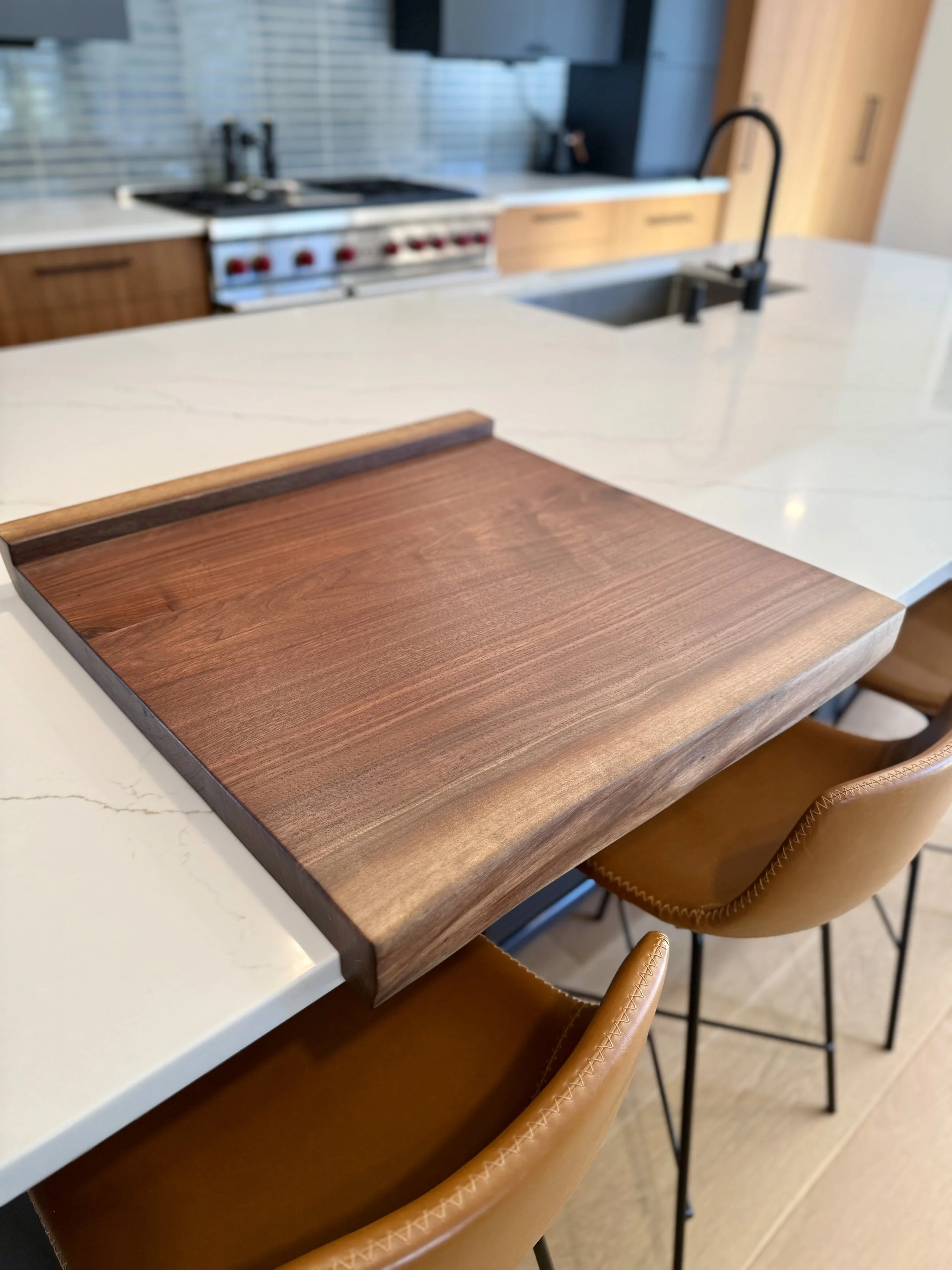 Close-up of a kitchen island with a walnut wooden serving tray on a white marble countertop, with bar stools underneath, and a stove, sink, and cabinets in the background.
