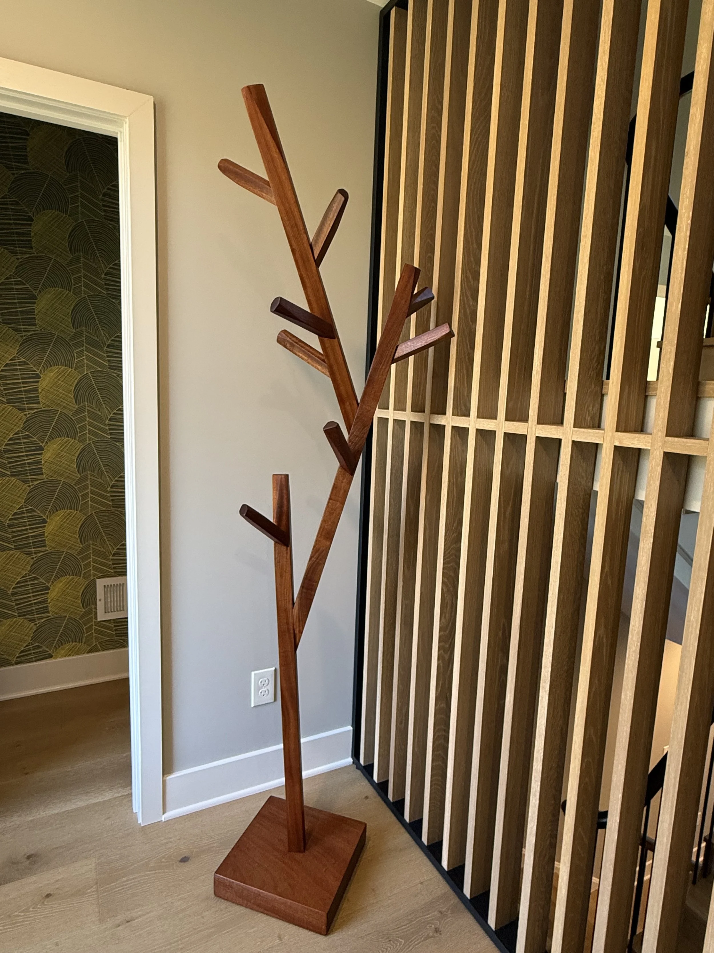Custom designed wooden coat rack designed like a tree with multiple branches, made from solid mahogany positioned near a wall and a wooden slat room divider in a modern interior.