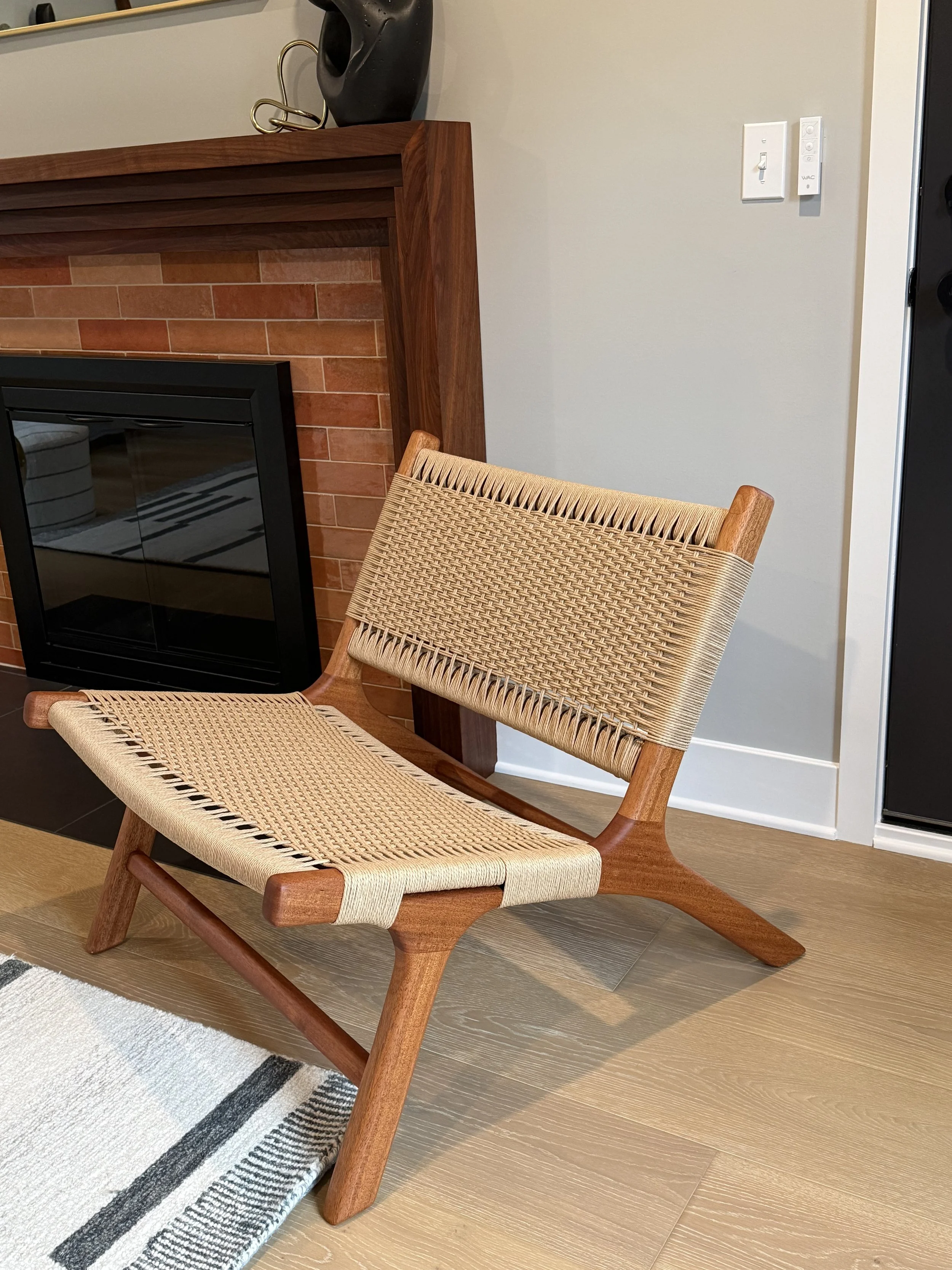 A wooden chair with woven seat and backrest in front of a fireplace with a brick surround and a wooden mantle. A decorative black object and a metallic sculpture are on the mantle. Part of a white area rug with black stripes is visible on the wooden 