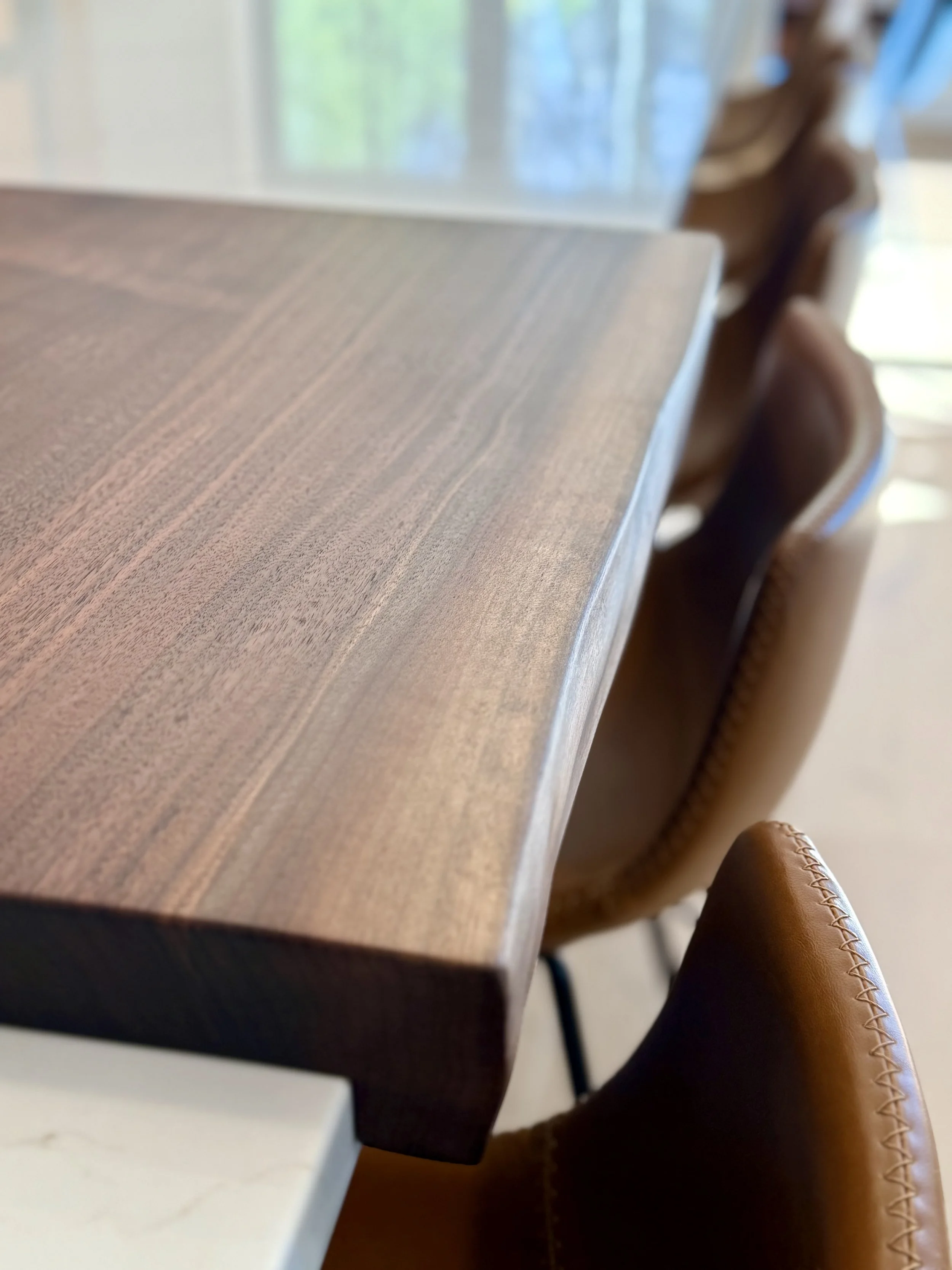 Close-up of a wooden table with a leather chair next to it in a well-lit room with windows in the background.