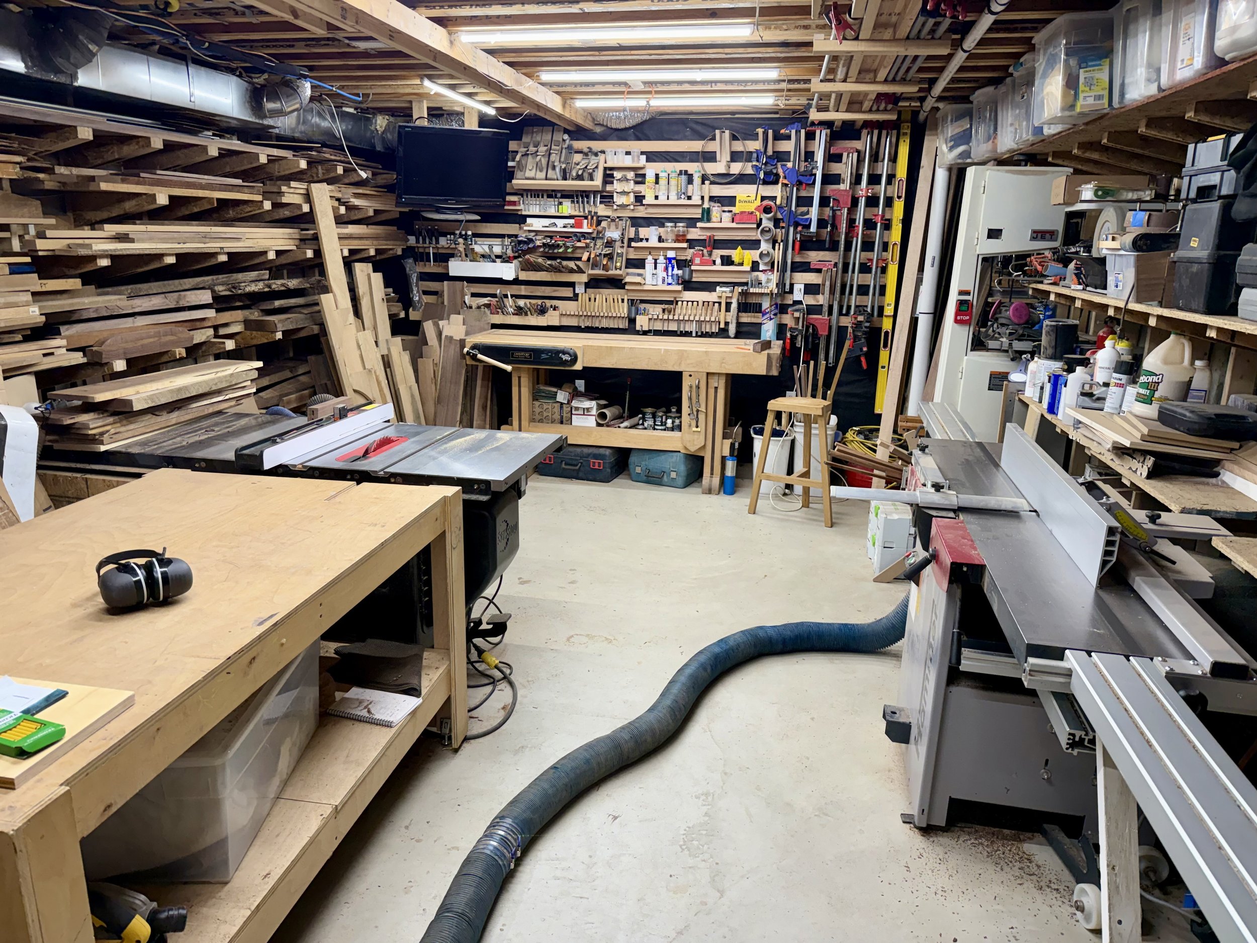 A cluttered woodworking workshop with shelves filled with tools, supplies, and wood pieces. Various power tools, a saw, and a yellow level are visible, along with a TV mounted on the wall and a large vacuum hose on the floor.