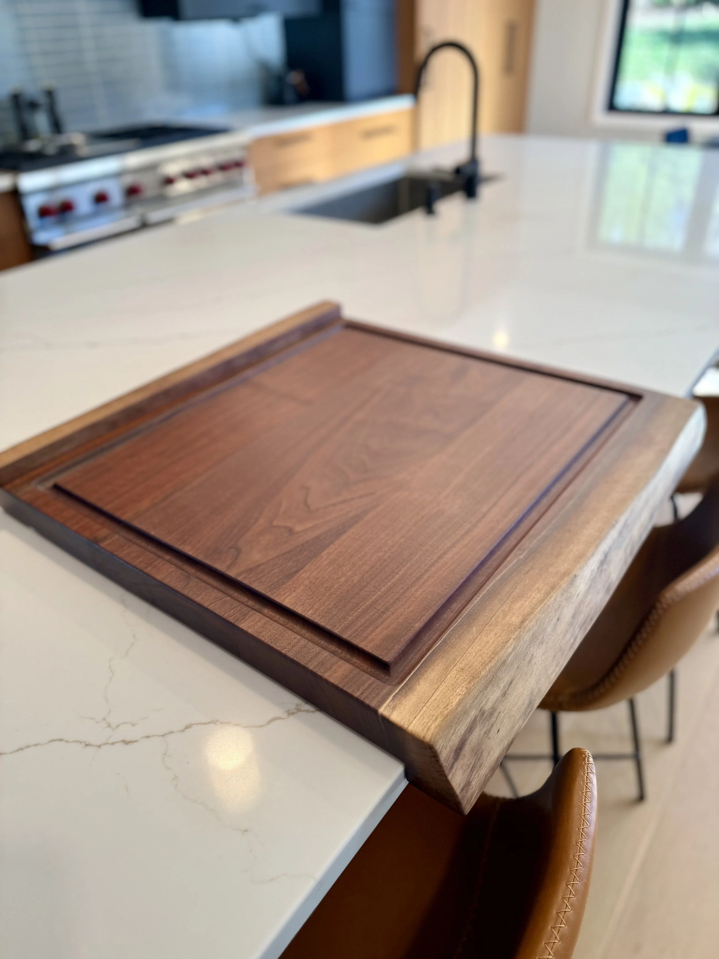 A custom designed walnut wood cutting board on a white marble kitchen island with brown leather chairs nearby.