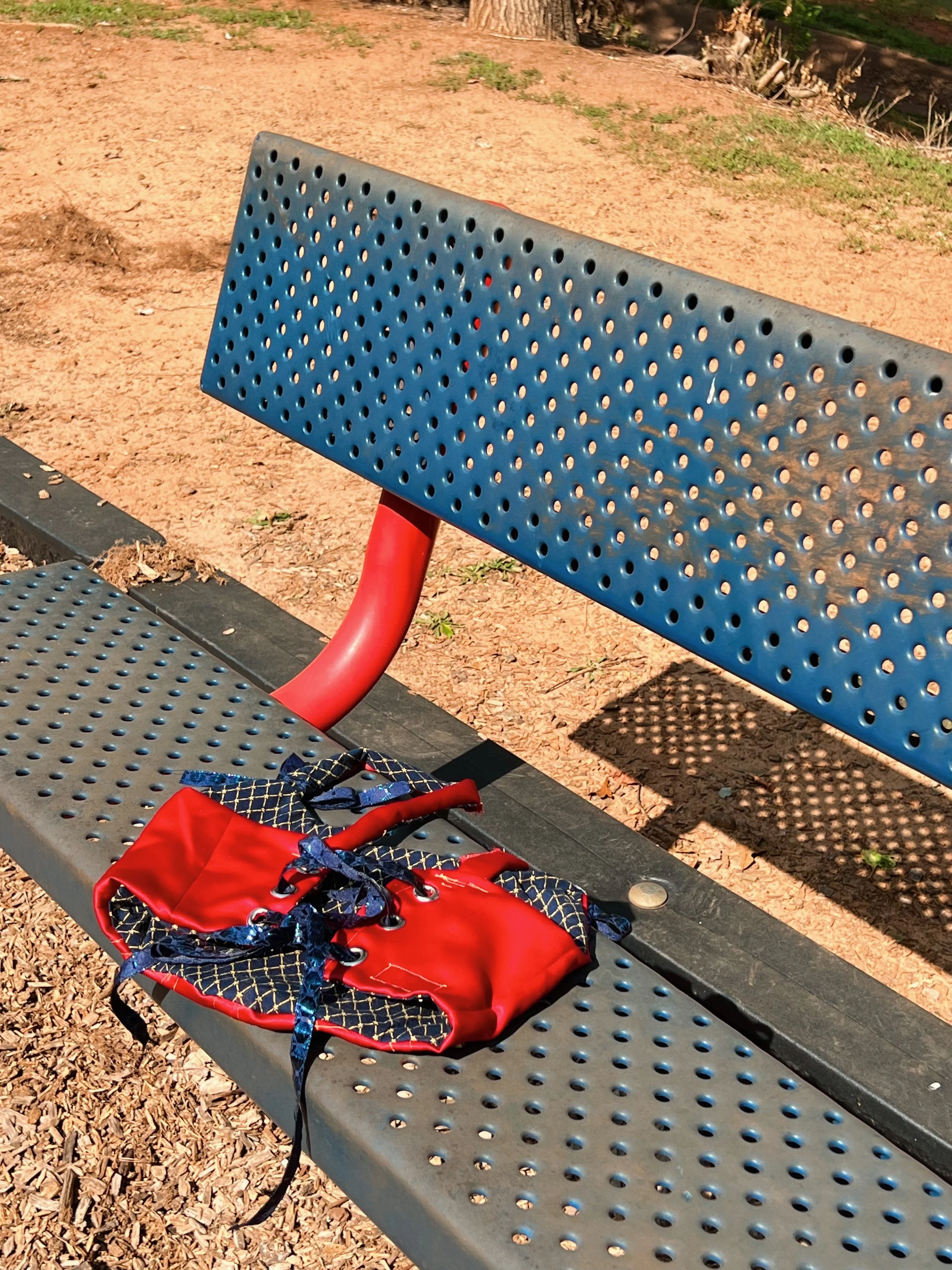 A blue perforated metal park bench with red metal legs, placed on a patch of dirt and grass. On the bench, there is a small red backpack with blue and black straps.