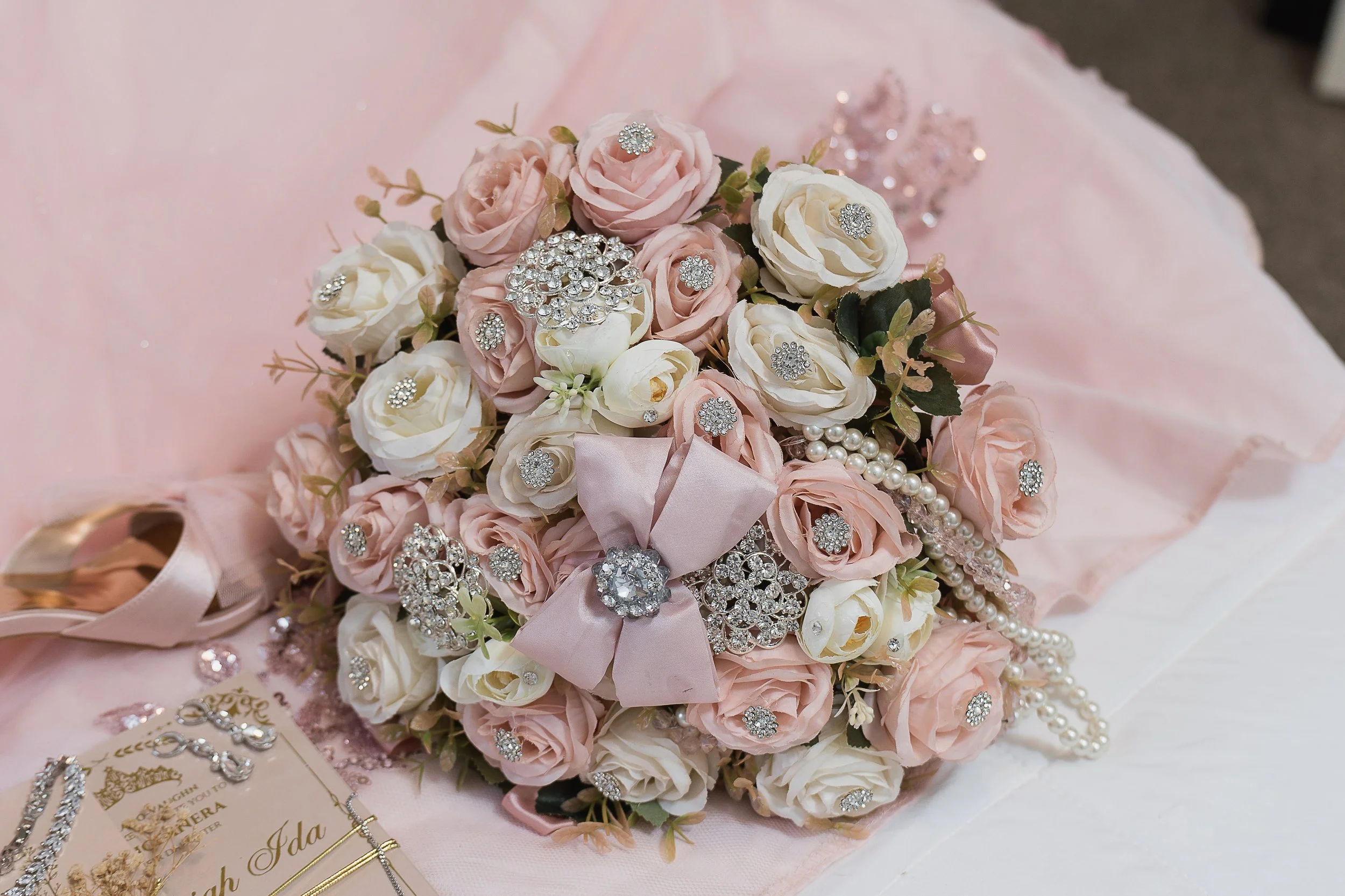 A bouquet of pink and white roses decorated with rhinestones and pearl jewelry, placed on a pink and white fabric with jewelry and a card nearby.
