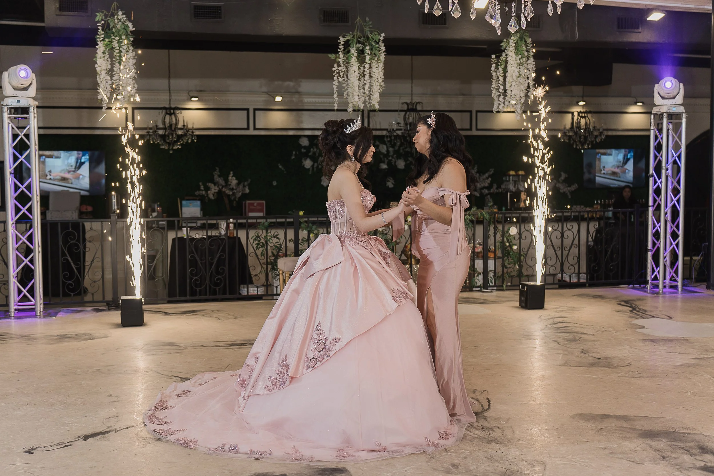 Mother and daughter holding hands and dancing at a celebration, with sparklers and decorative lighting in the background.