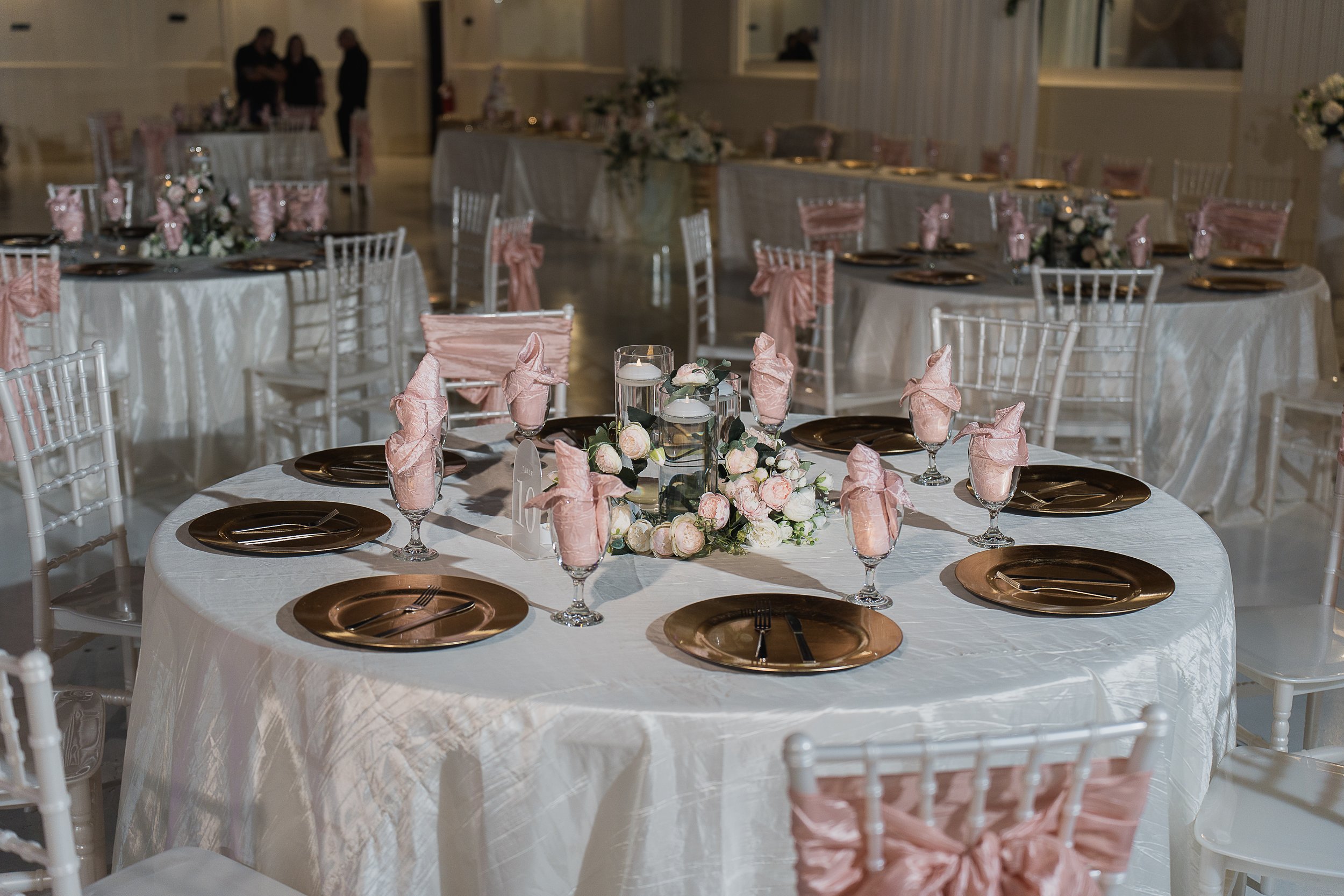 Elegant banquet hall decorated for a Quinceañera with round tables adorned with white tablecloths, pink napkins, gold plates, and floral centerpieces featuring pink and white roses.