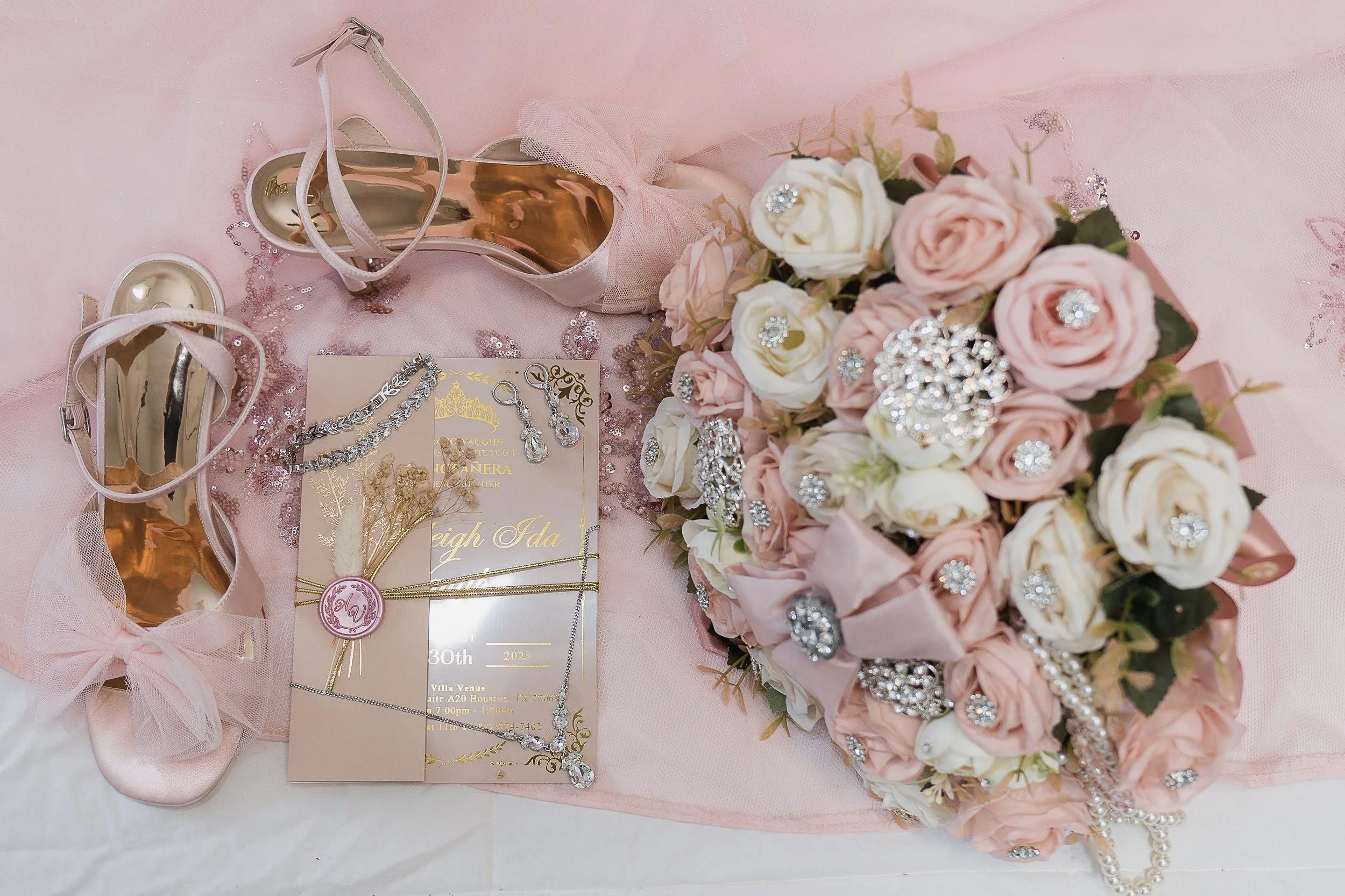 Pink satin and tulle bridal shoes, a wedding invitation with jewelry and a bridal bouquet of pink and white roses with rhinestone embellishments, all arranged on a pink tulle fabric.