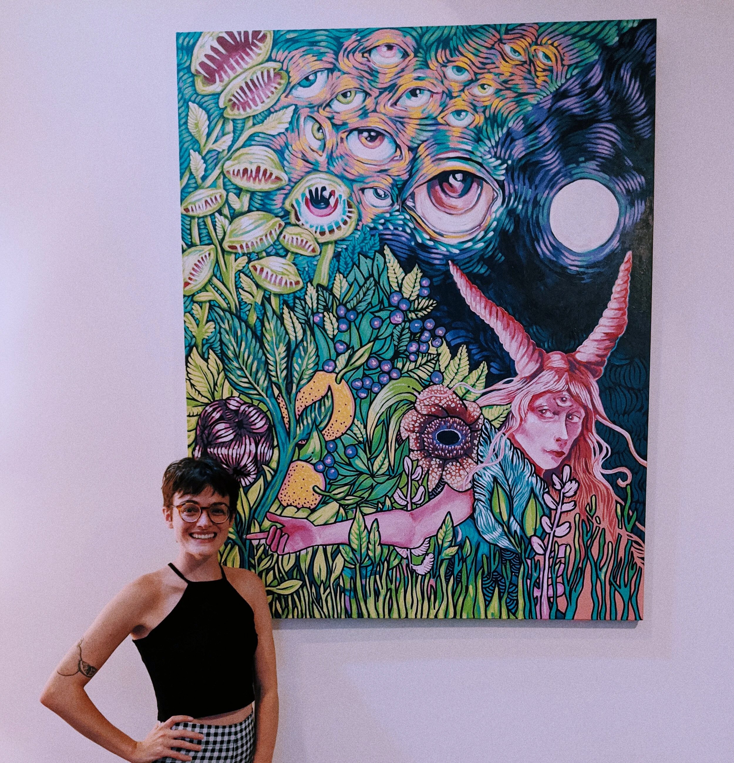 A person with short dark hair, glasses, and a sleeveless black top standing in front of a colorful, surreal painting with multiple eyes, roses, and abstract patterns.