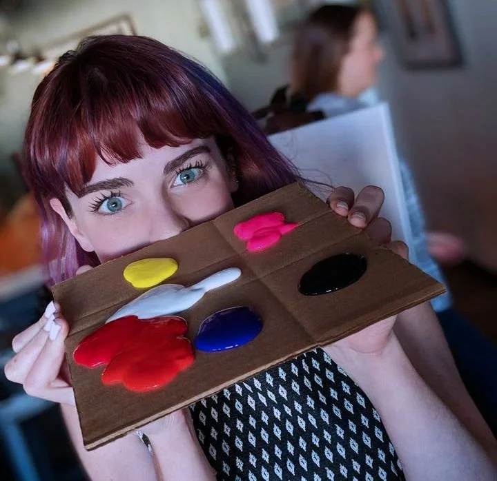 Young woman with red hair and blue eyes holding a wooden palette with colorful paint, including yellow, white, red, blue, black, and pink, in a room with other people in the background.