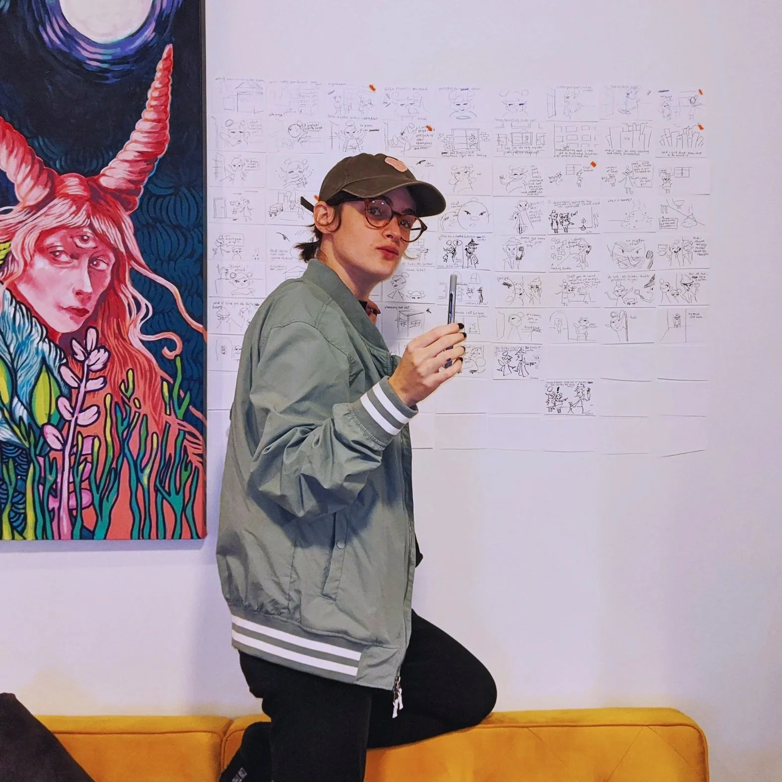 A person with glasses and a cap, kneeling on a yellow bench in front of a wall with drawings and a large colorful painting of a woman with a unicorn horn. The person is holding a pen and looking at the camera.