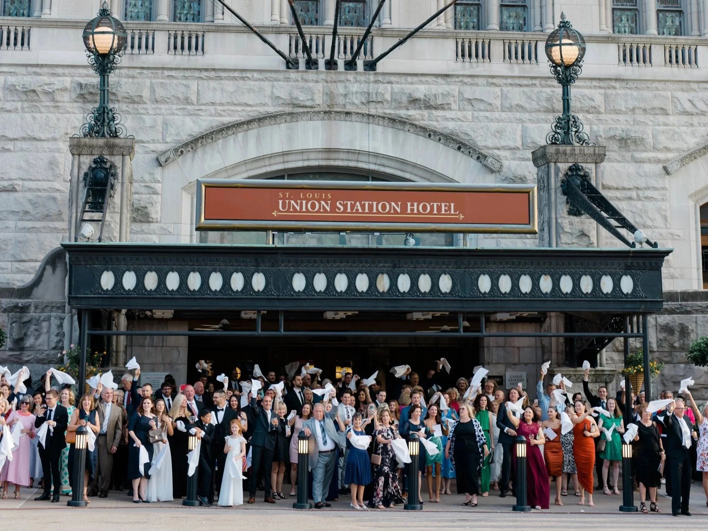 A large group of people dressed in formal attire gathered outside the St. Louis Union Station Hotel, celebrating with some waving white cloths in the air.