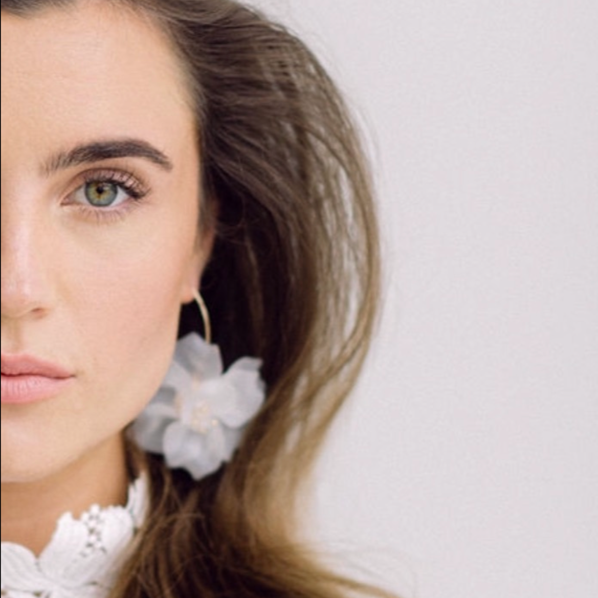 Close-up of a woman's face, showing her eye, eyebrow, and part of her lips, with brown hair, an earring, and a white flower accessory.