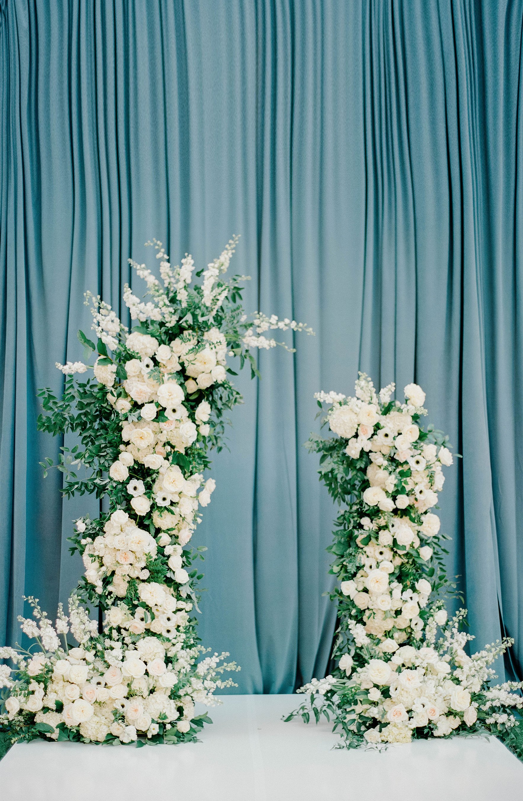 Two tall floral arrangements featuring white roses and greenery set against a blue curtain backdrop.