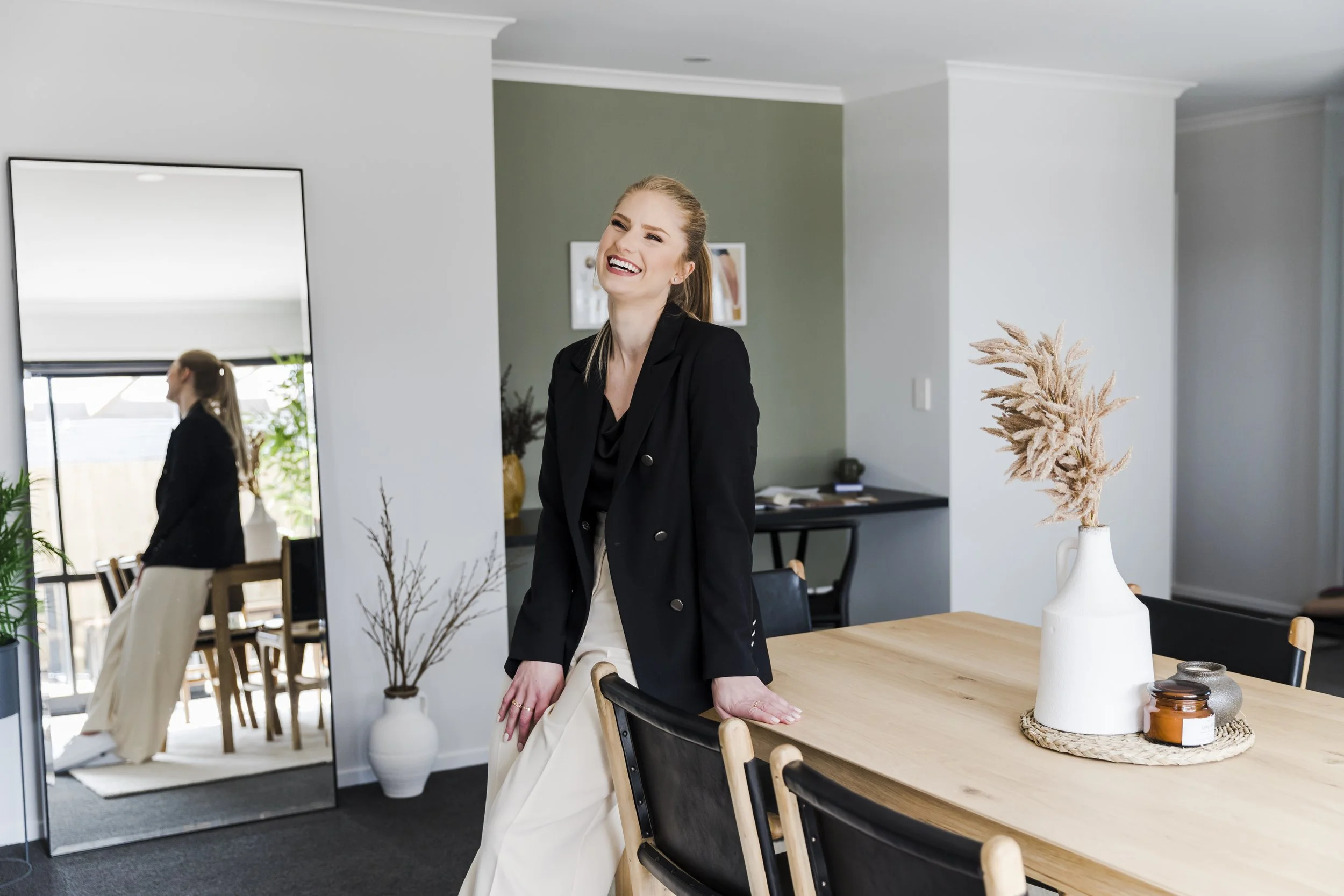 A woman with blonde hair in a ponytail, wearing a black blazer and cream-colored pants, laughing and leaning on a wooden dining table in a modern, well-lit room. A large mirror reflects her side profile and a dining area with chairs and potted plants. The table has a white vase with dried beige pampas grass, a woven placemat, and small decorative jars.