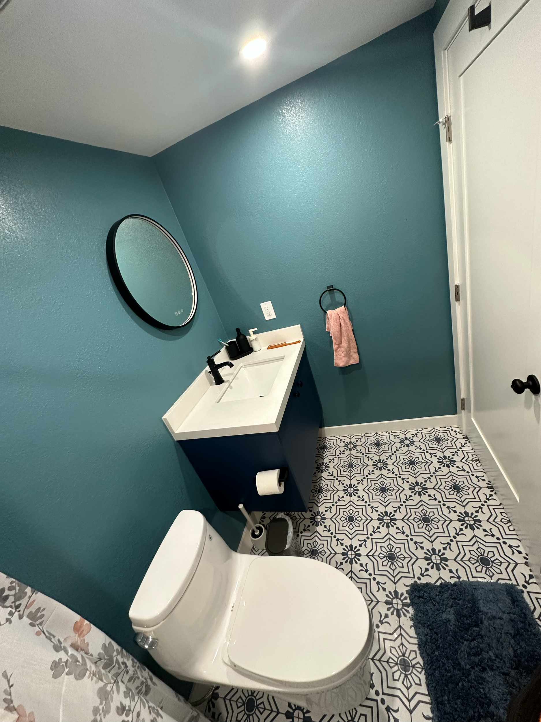 Modern bathroom with blue walls, white toilet, patterned floor tiles, a round mirror, and a blue vanity with a sink.