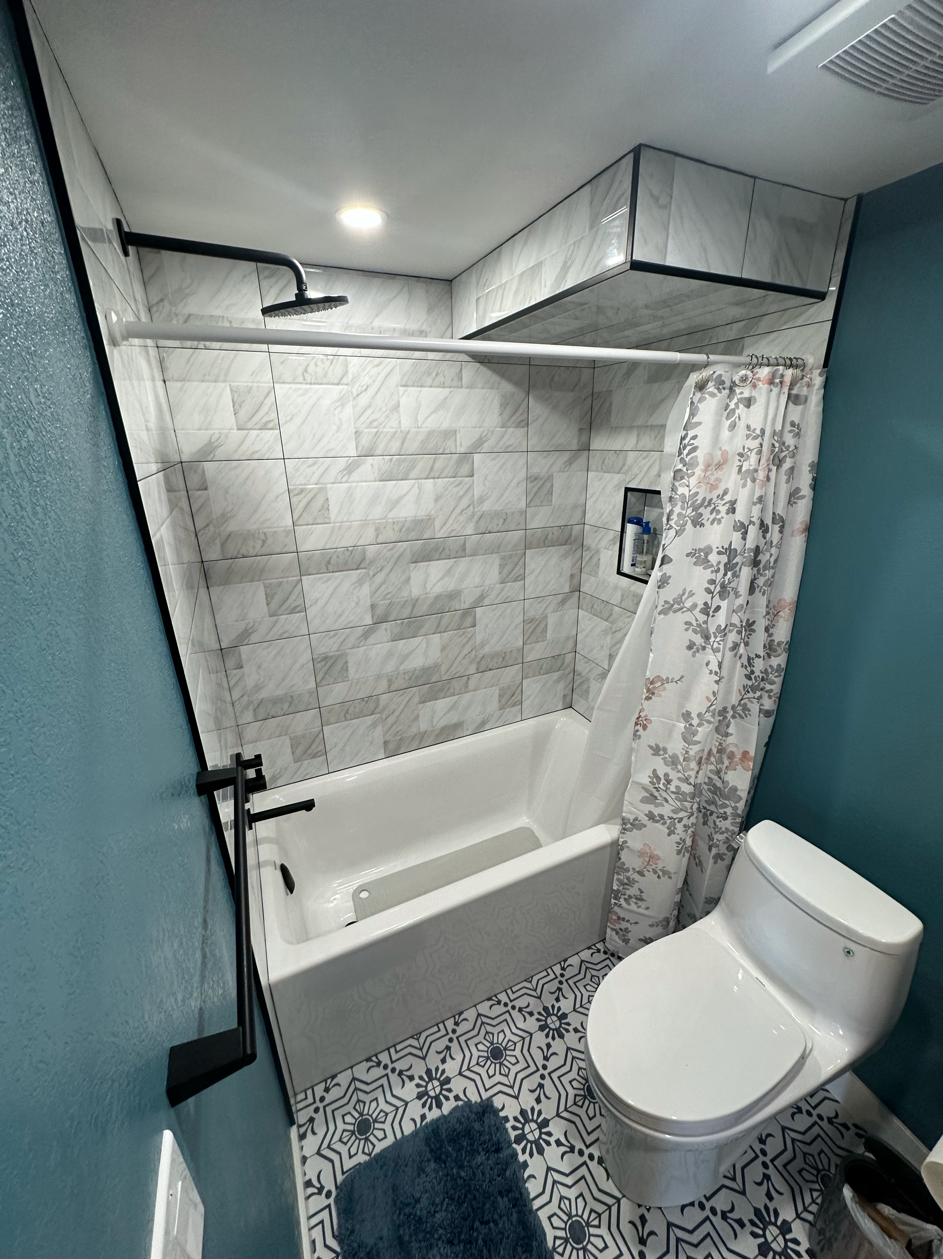 Bathroom featuring a white toilet, a bathtub with gray tile walls, floral shower curtain, black showerhead, and patterned floor tiles.