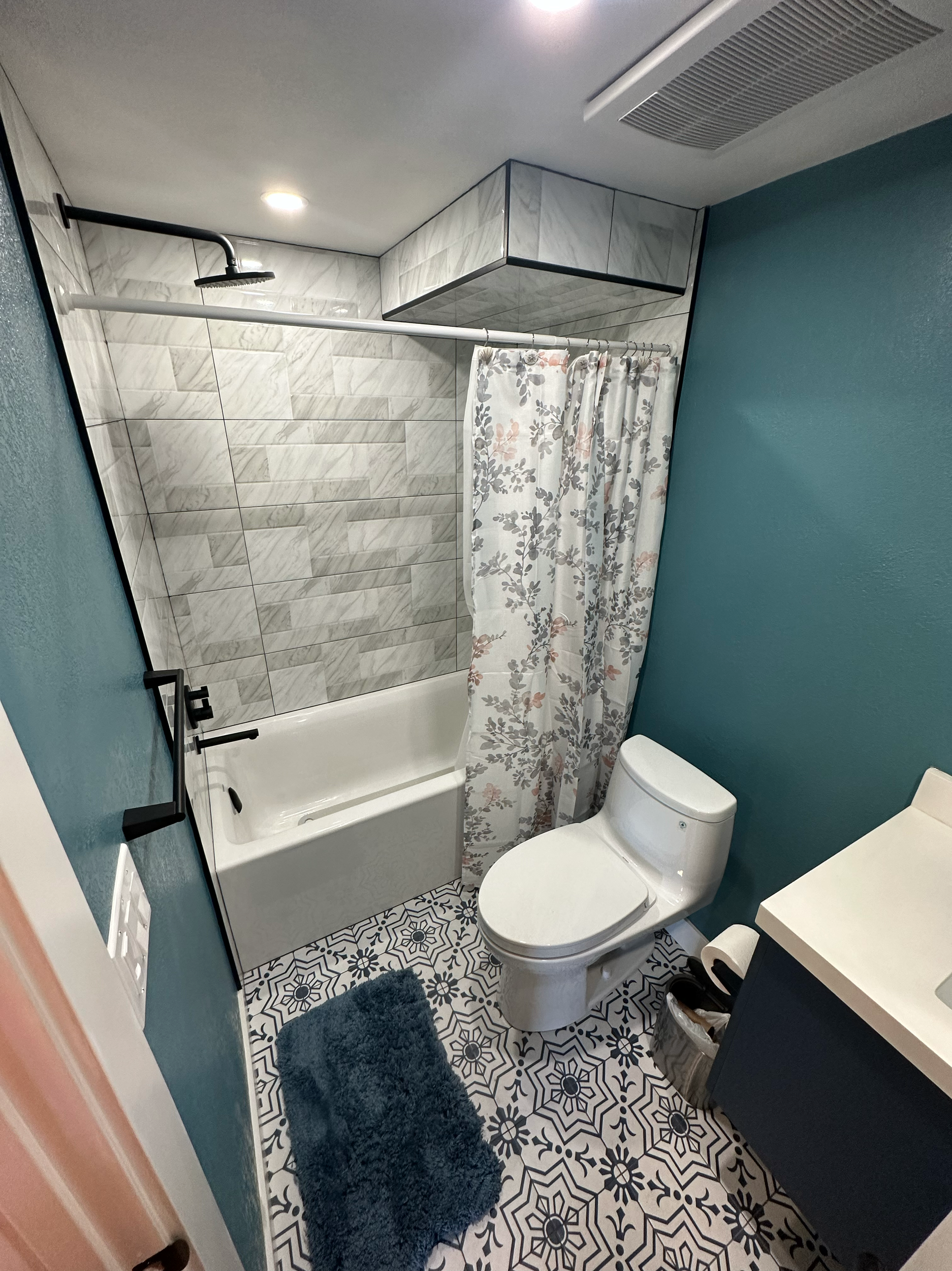 Bathroom with blue walls and patterned floor tiles, featuring a bathtub with a floral shower curtain, a toilet, and a vanity with a sink.