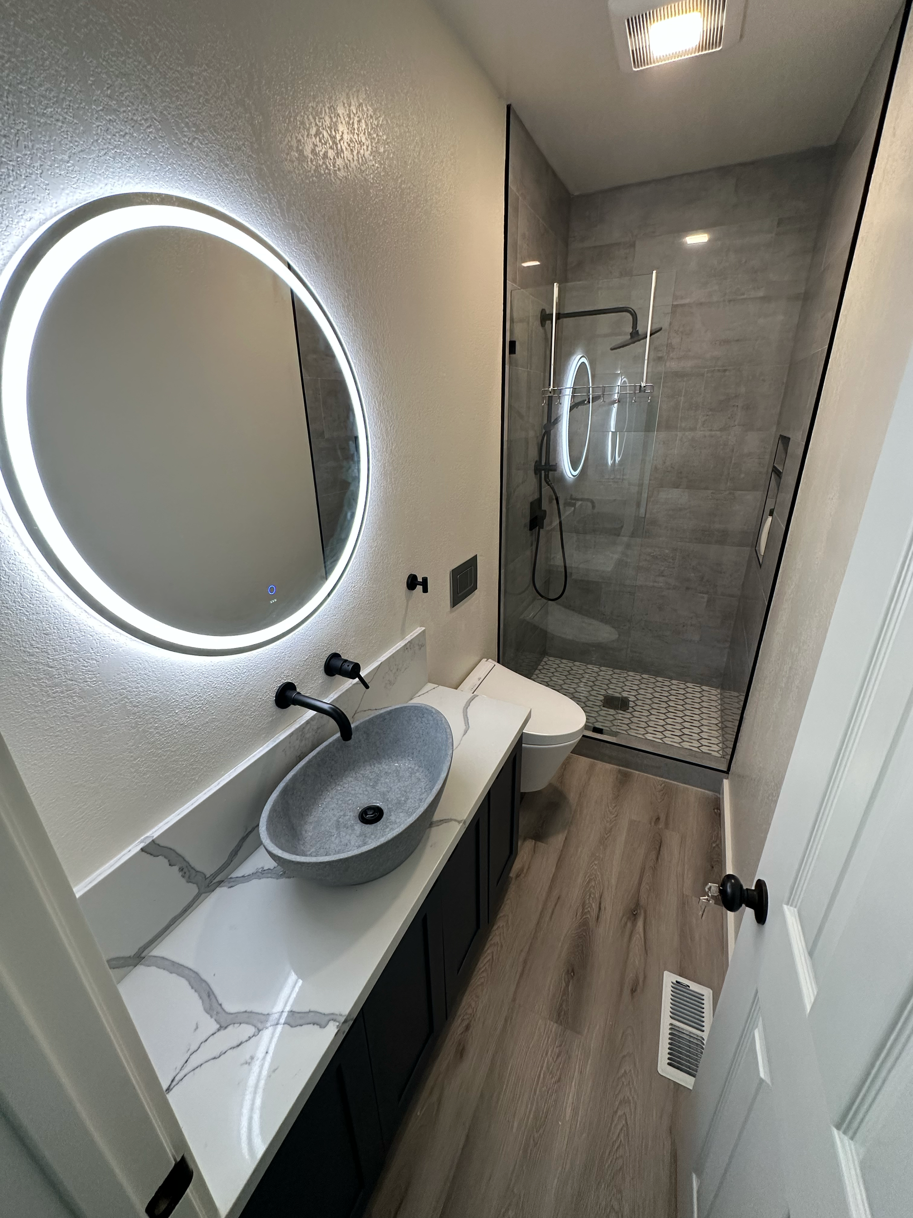 Modern bathroom with a wall-mounted circular mirror and LED lighting, a stone vessel sink on a white marble countertop, matte black fixtures, glass-enclosed shower with gray tile walls, and wood-look flooring.