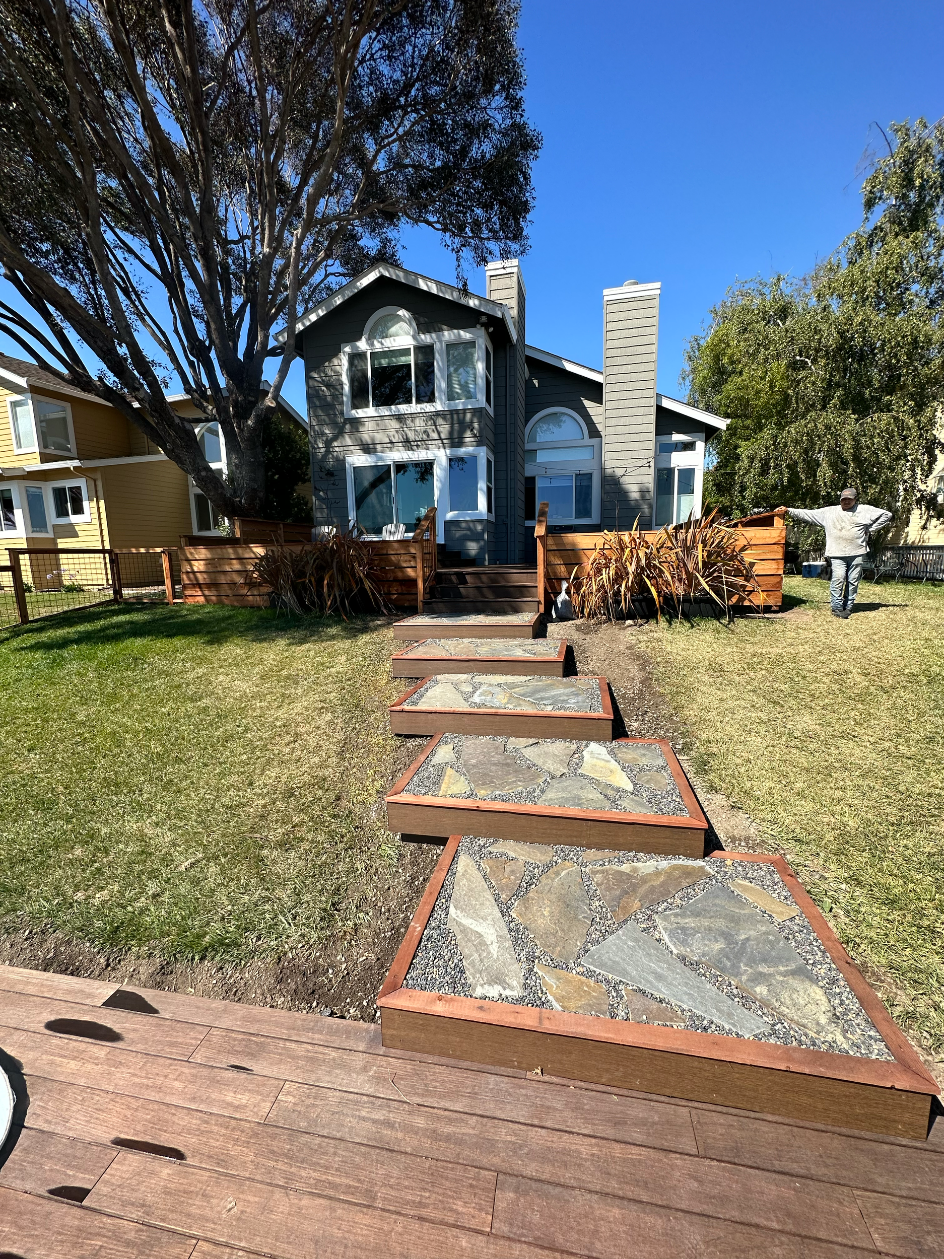 Backyard view with stone walkway leading to a house, trees on the left, and person standing in the background.
