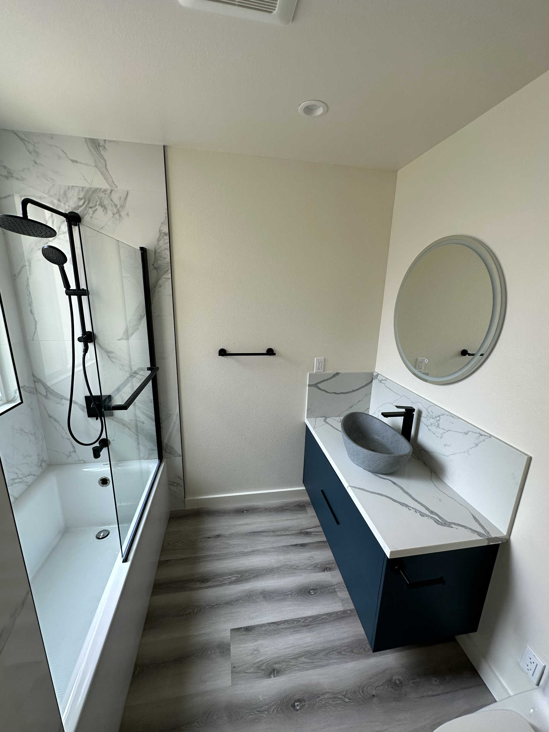 Modern bathroom with circular mirror, marble countertop, vessel sink, black hardware, glass shower enclosure, and wood-look flooring.