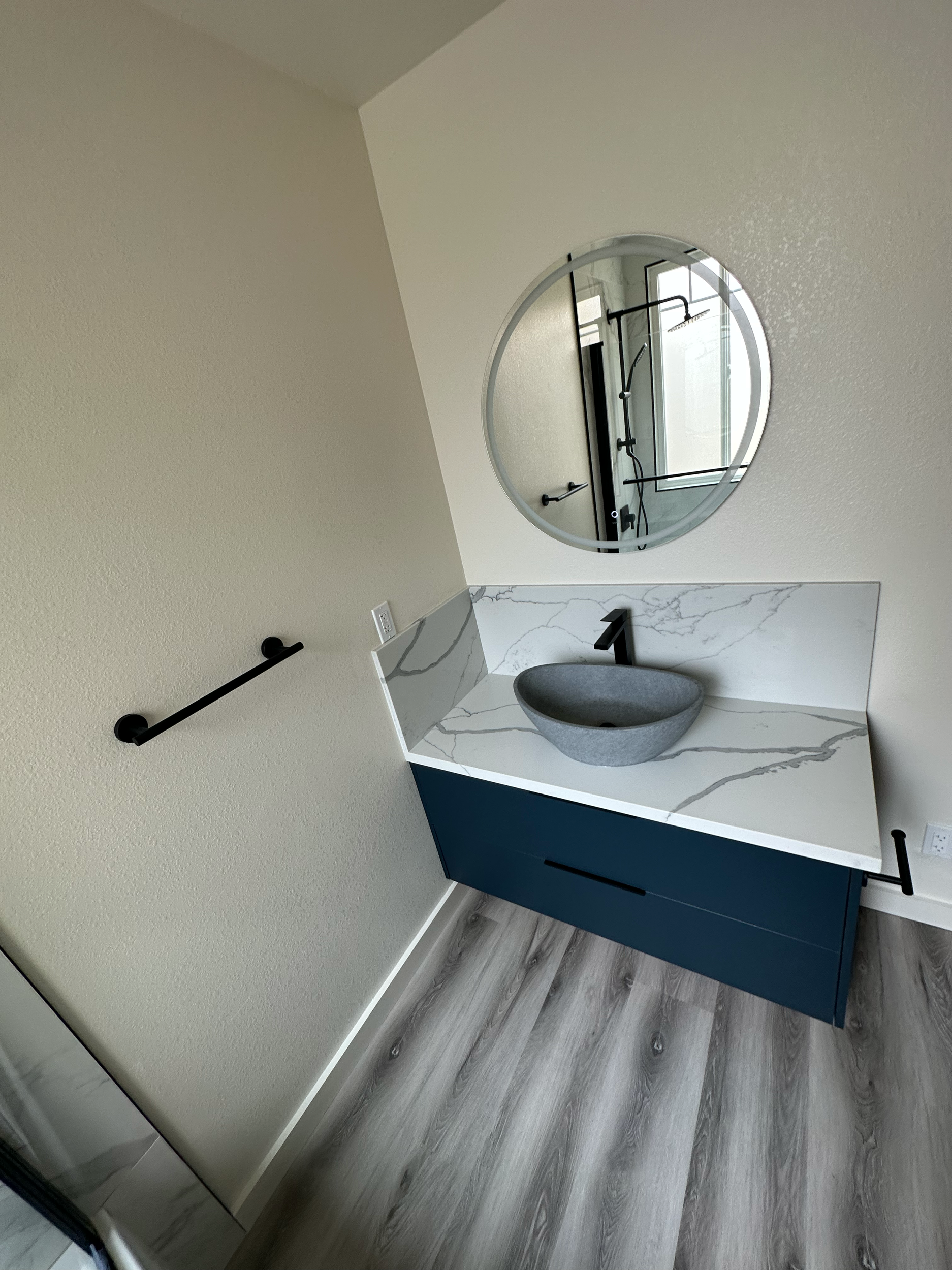 Modern bathroom with round mirror, vessel sink, marble countertop, and dark cabinetry on light wood flooring.