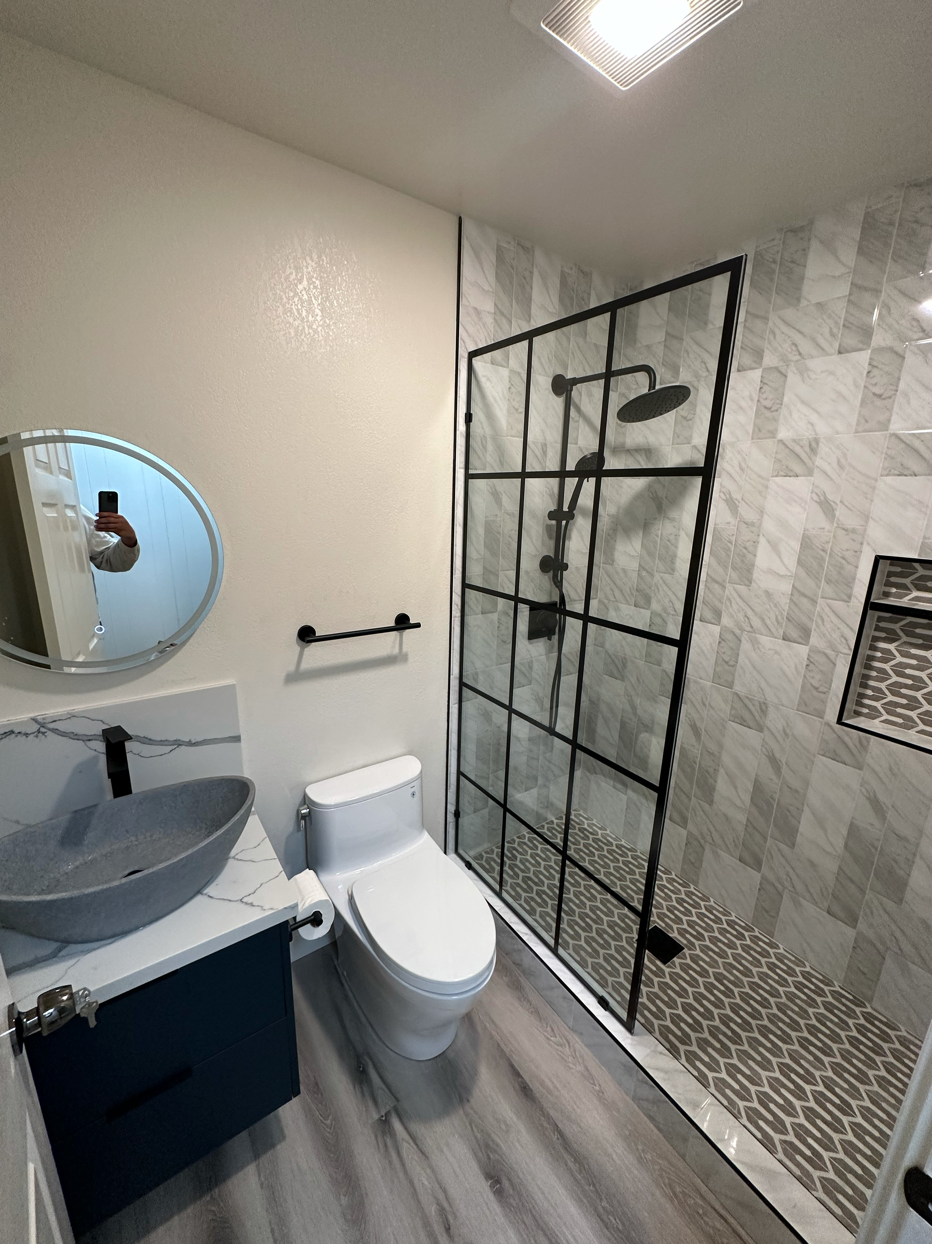 Modern bathroom with marble-tiled shower, glass door, round mirror, vessel sink, and toilet on wooden floor.