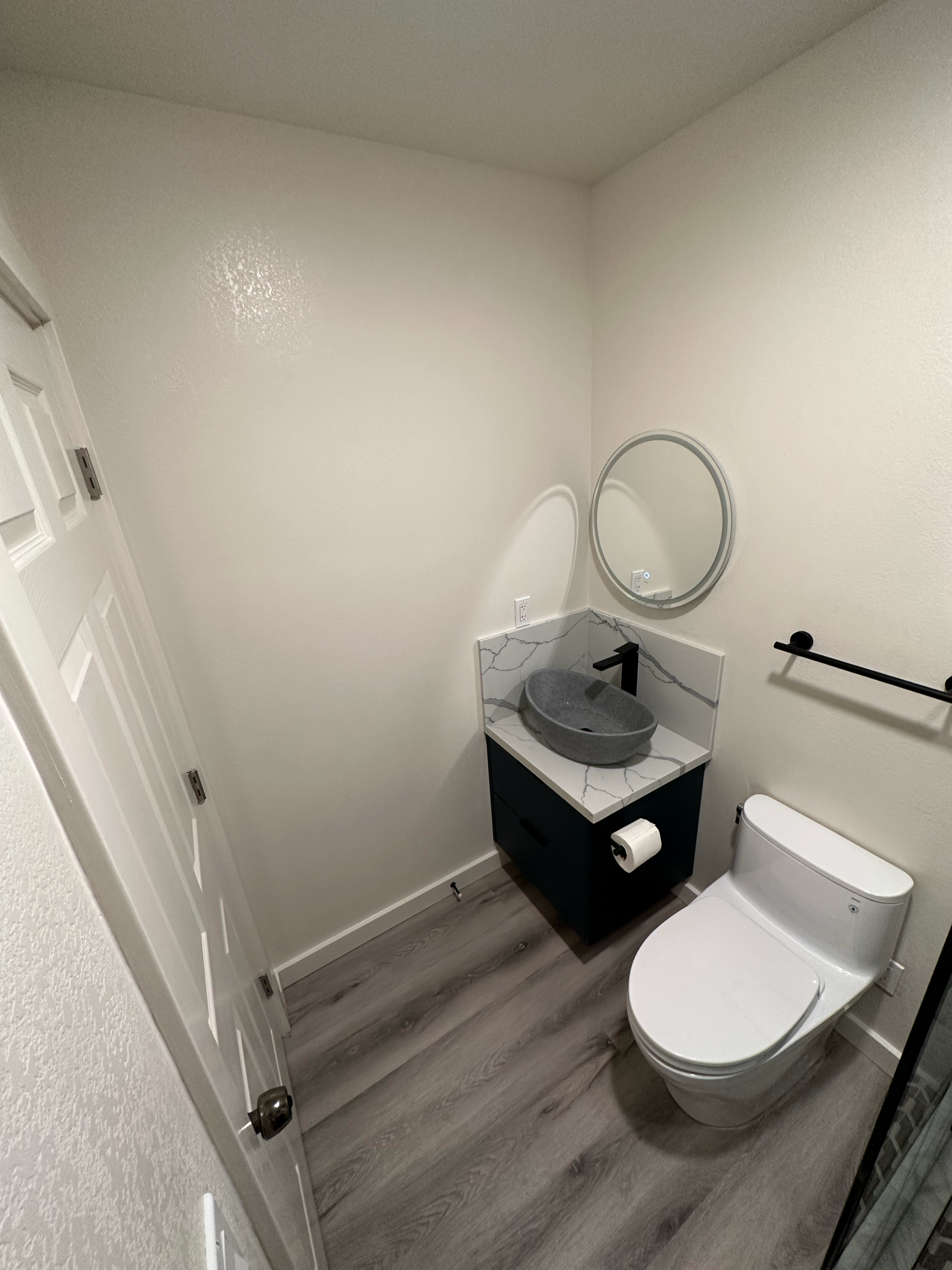 Modern bathroom with a round mirror, vessel sink, and toilet, featuring light wood flooring and white walls.