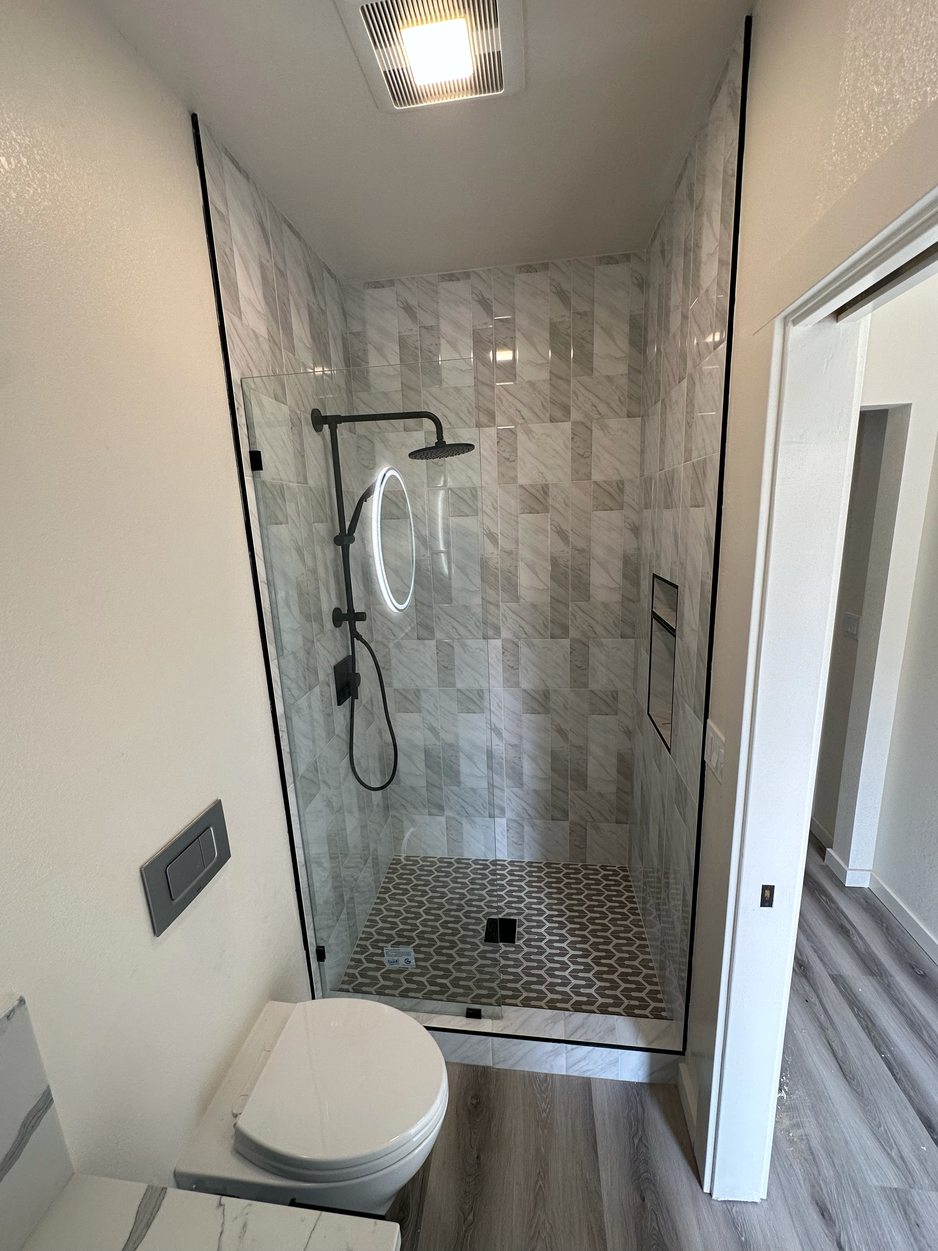 Modern bathroom with glass shower enclosure, marble tile walls, circular mirror with LED lighting, and wooden flooring.