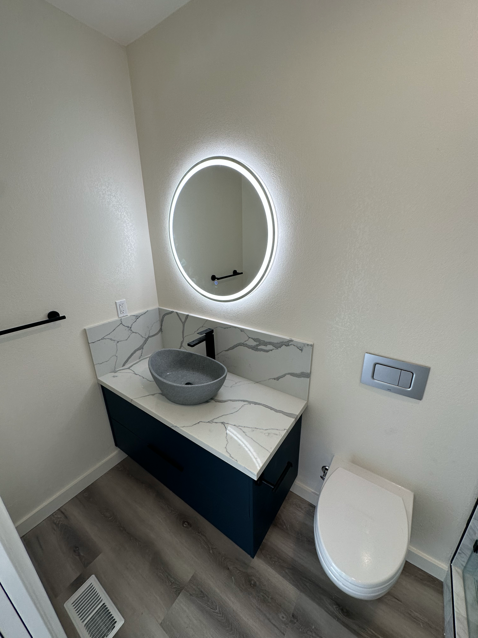 Modern bathroom with a circular lighted mirror, a vessel sink on a marble countertop, a wall-hung toilet, and wood-style flooring.