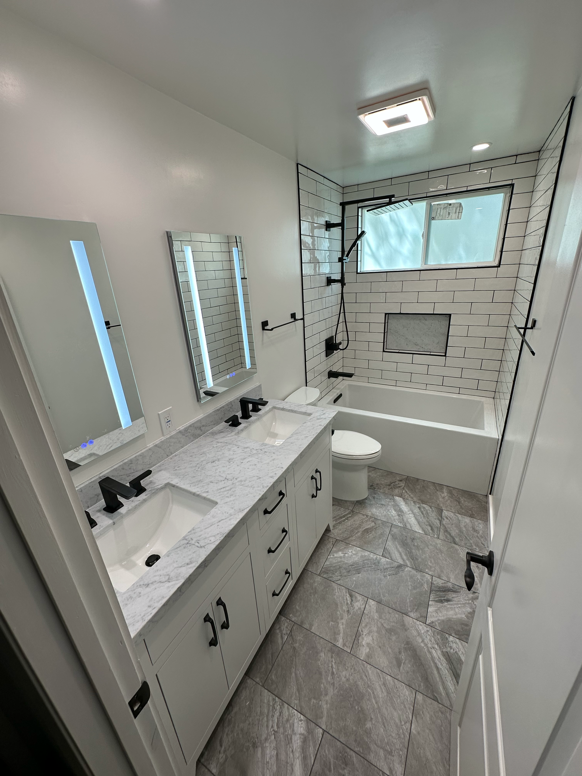 Modern bathroom with white tile shower, double vanity with marble countertop, black fixtures, large mirror, and gray tile floor.