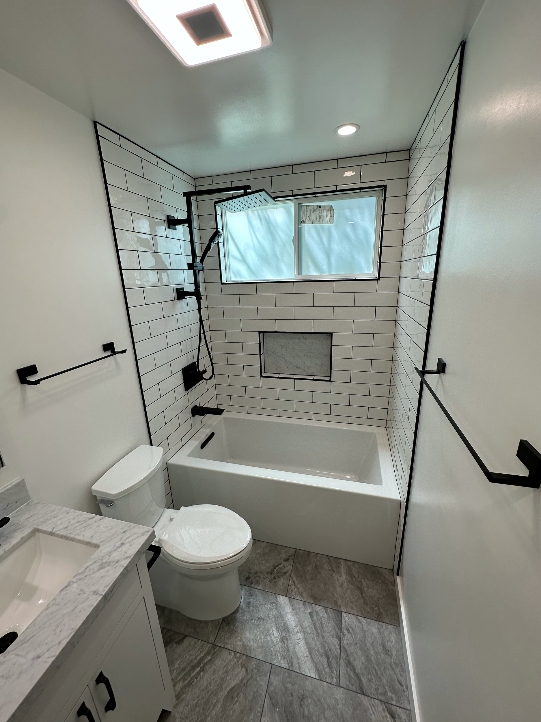 Modern bathroom with black fixtures, white subway tiles, a bathtub, a toilet, and a black-framed window.