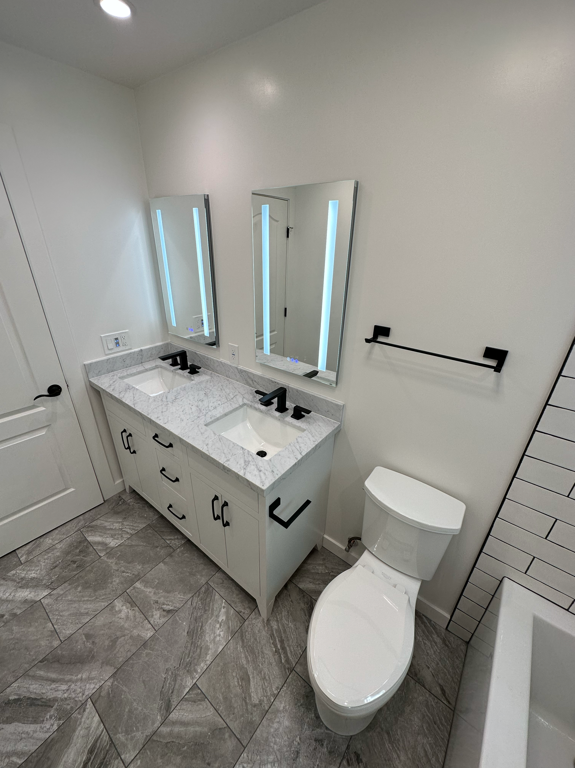 Modern bathroom featuring a vanity with a marble countertop, undermount sink, dual lighted mirrors, and faucet with black handles. There is also a toilet, with a tiled floor and wall in gray tones.