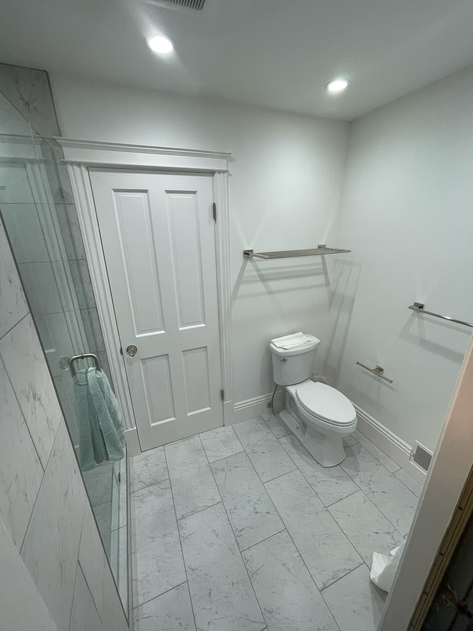 Modern bathroom with white tiled floor, toilet, white door, glass shower, and towel rack.