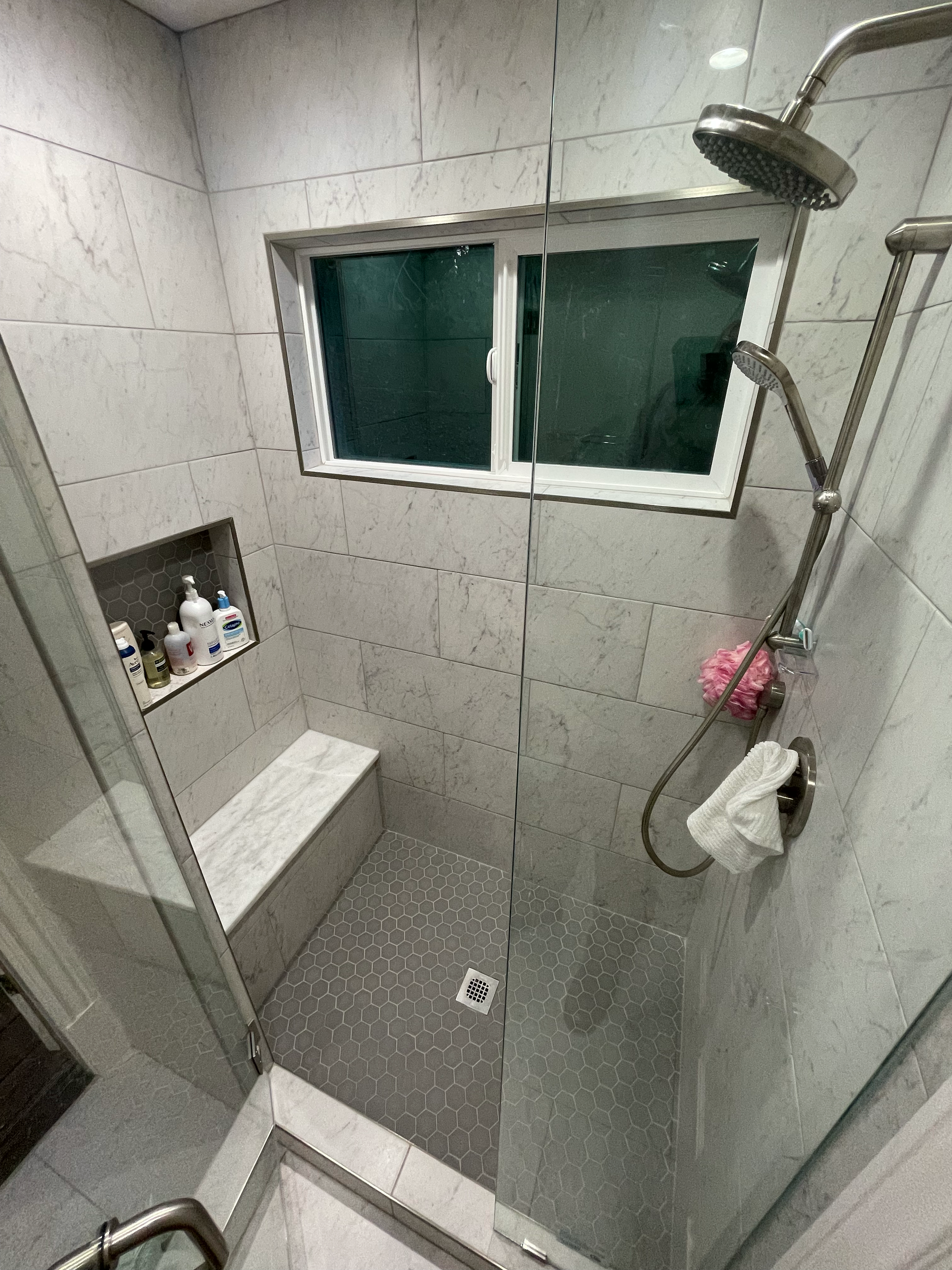 Modern tiled shower with showerheads, window, bench, and toiletries in a wall niche.