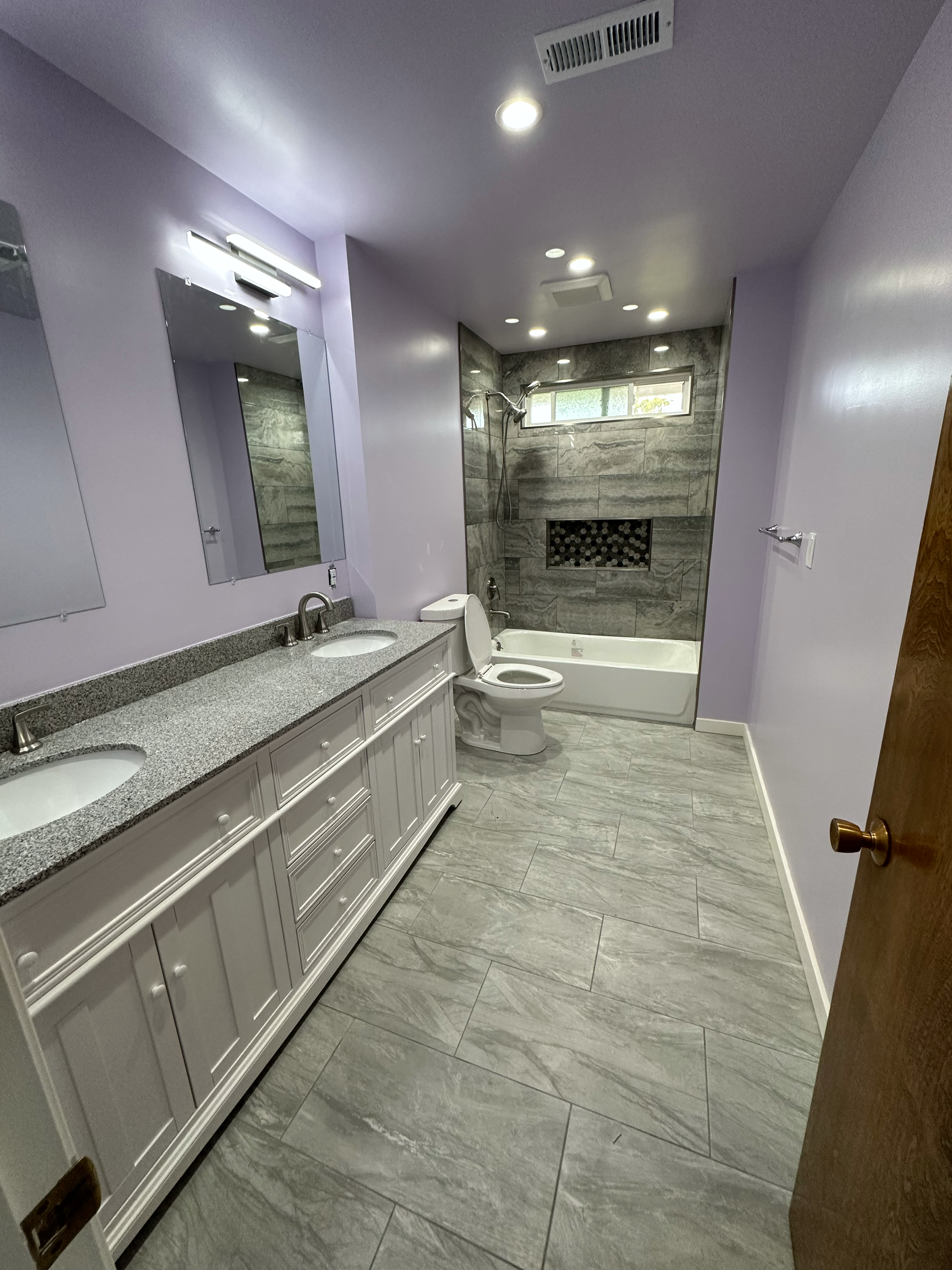 Modern bathroom with light purple walls, marble tiled floor, double vanity with gray countertop, large mirror, walk-in shower with natural stone accent, and white bathtub.