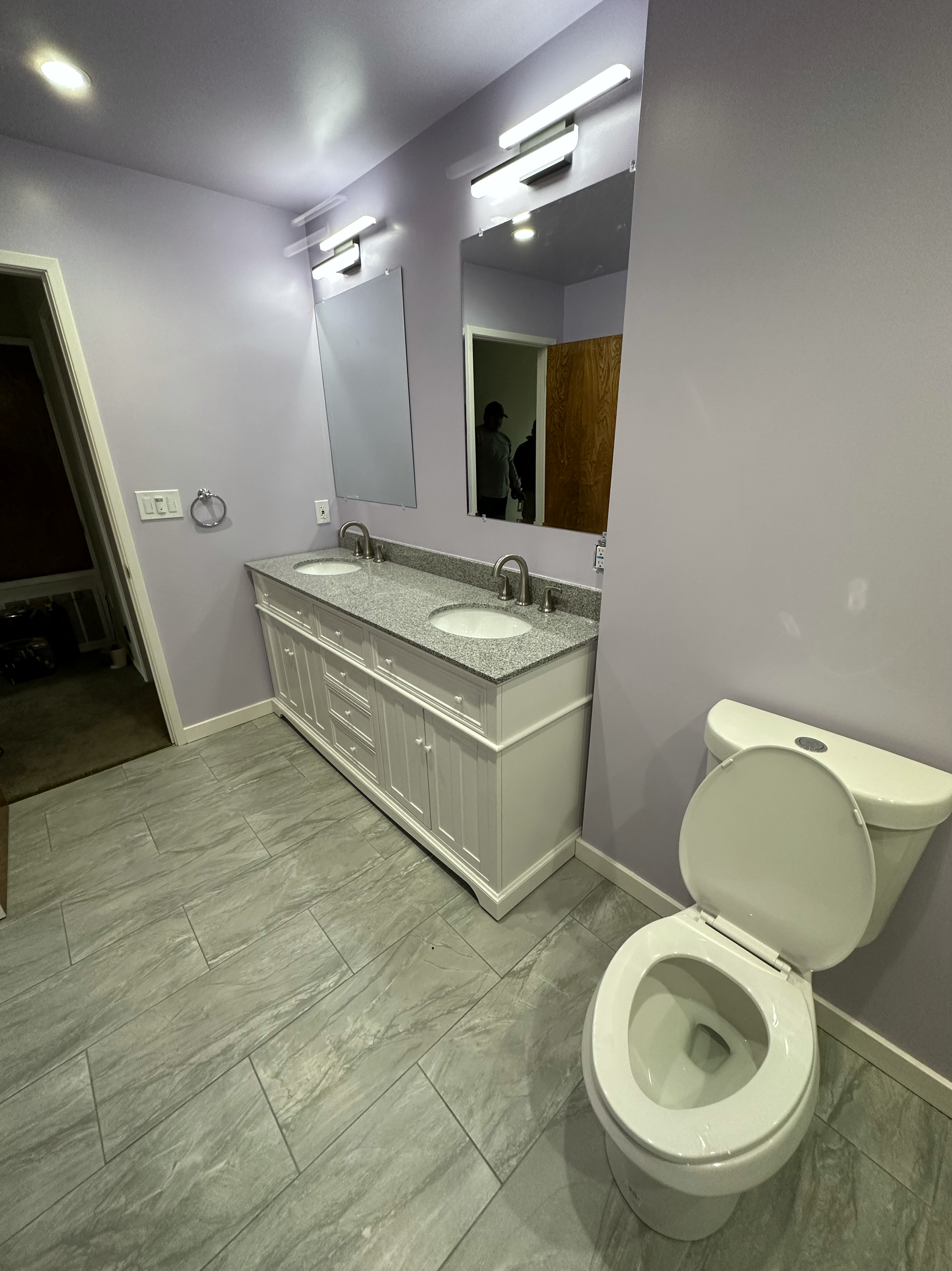 Bathroom with purple walls, gray tile floor, white vanity with granite countertop, double sinks and mirrors, and a toilet.